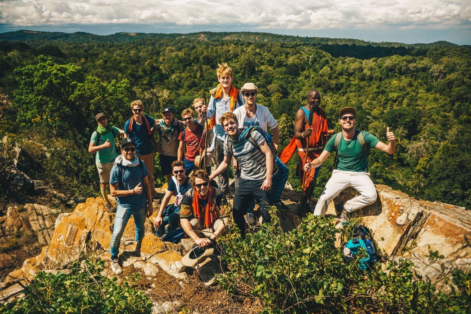 Een groep reizigers poseert en lacht samen op een rotsachtige heuveltop in Afrika, omringd door weelderige groene bossen en heuvels in de verte onder een gedeeltelijk bewolkte hemel.