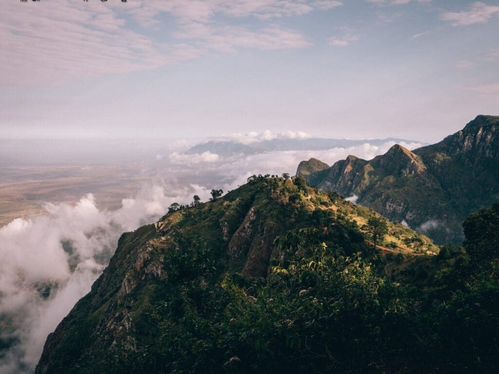 A lush, green mountain ridge in Tanzania rises above the clouds under a partly cloudy sky, with distant valleys and peaks visible on the horizon—an unforgettable sight for anyone travelling through Africa.