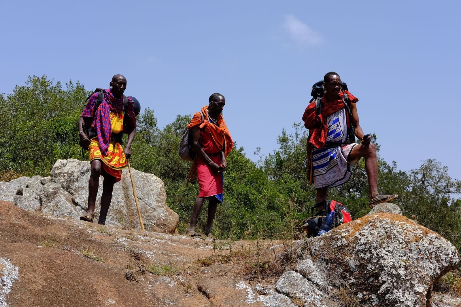 Three Maasai men in traditional clothing stand on rocky terrain outdoors in Kenya, surrounded by greenery under a clear blue sky. One man leans on a walking stick, another stands upright, and the third rests his foot on a rock—capturing the spirit of Africa.