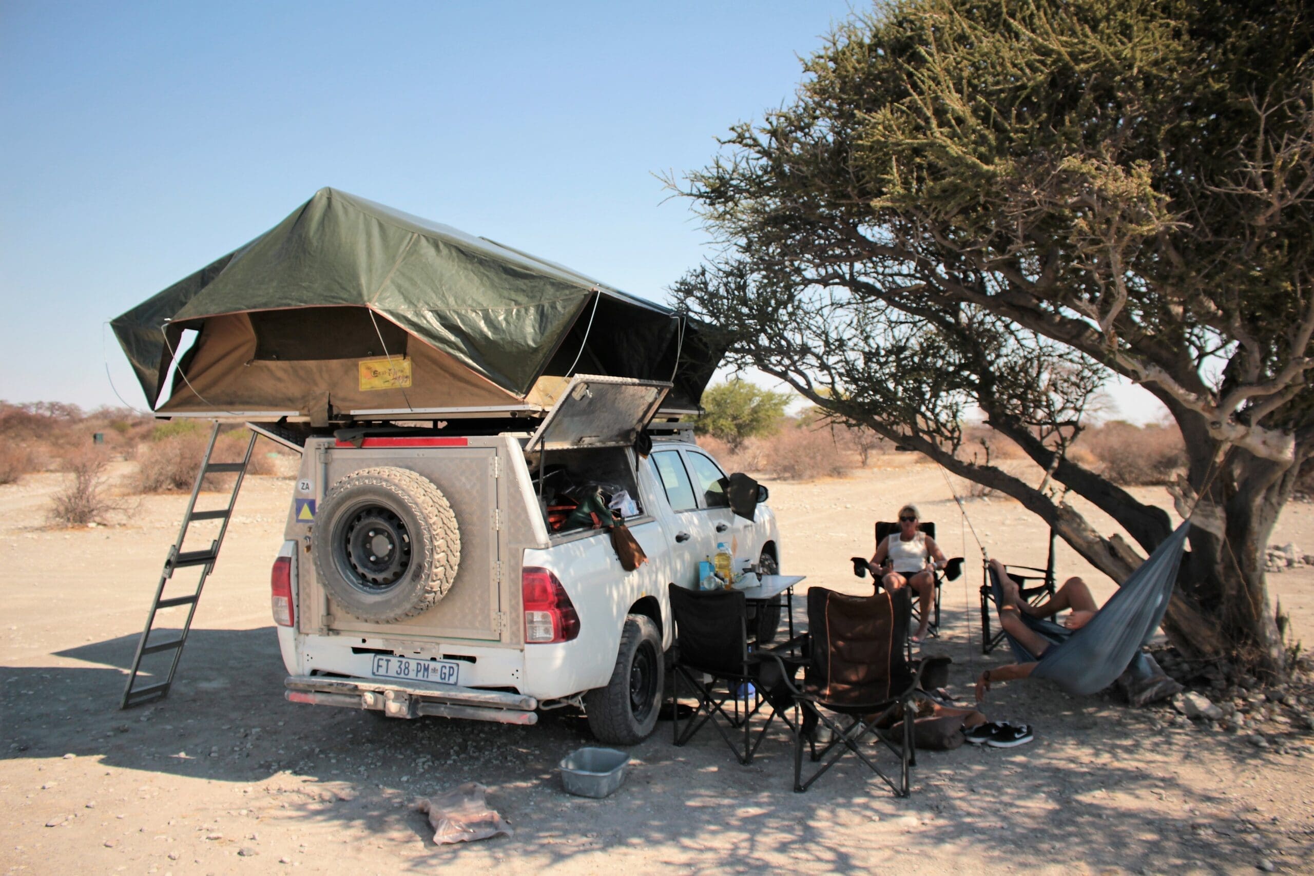 Een witte SUV met een daktent staat geparkeerd naast een boom in een droog, open landschap. Er omheen staan stoelen en kampeerspullen. Twee mensen relaxen in de schaduw; één zit in een hangmat die aan de boom hangt.