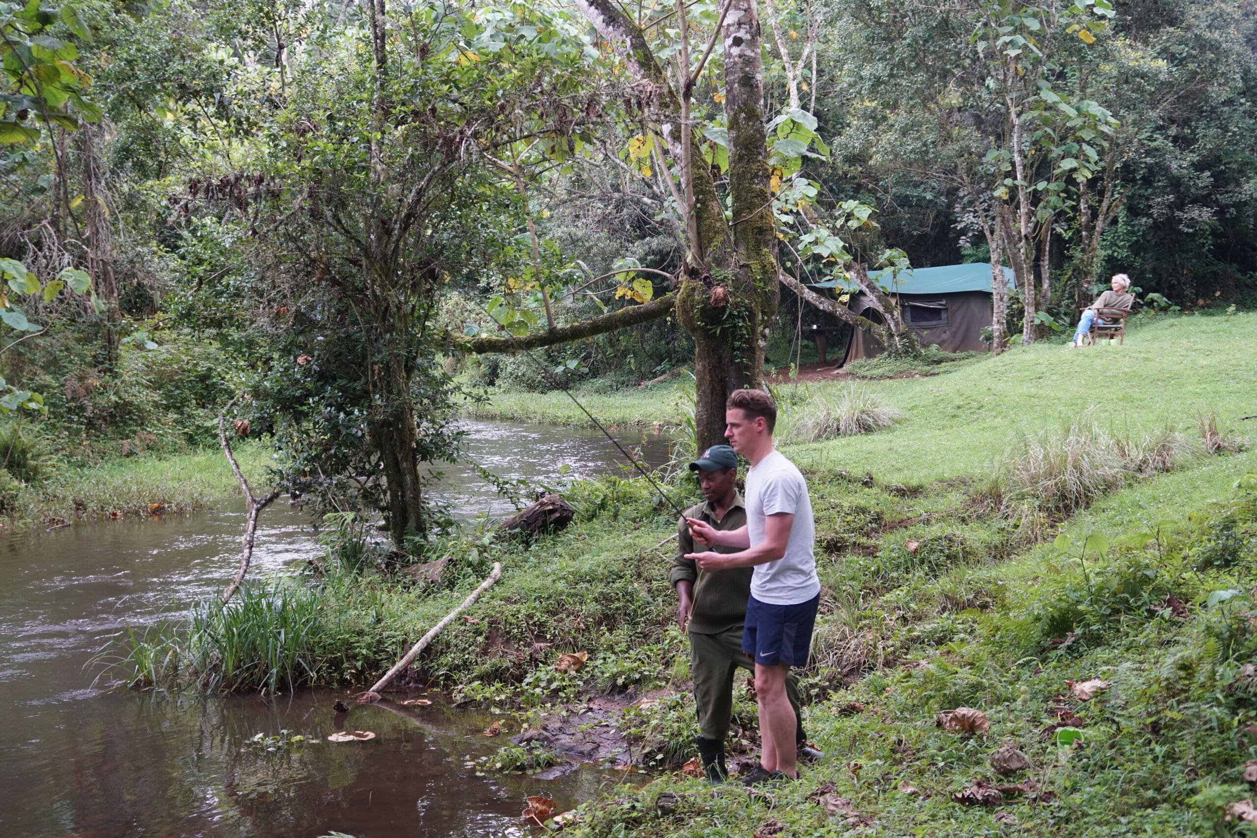 Man en gids vissen samen langs een rivier in de groene hooglanden van Kenia met tentenkamp op de achtergrond