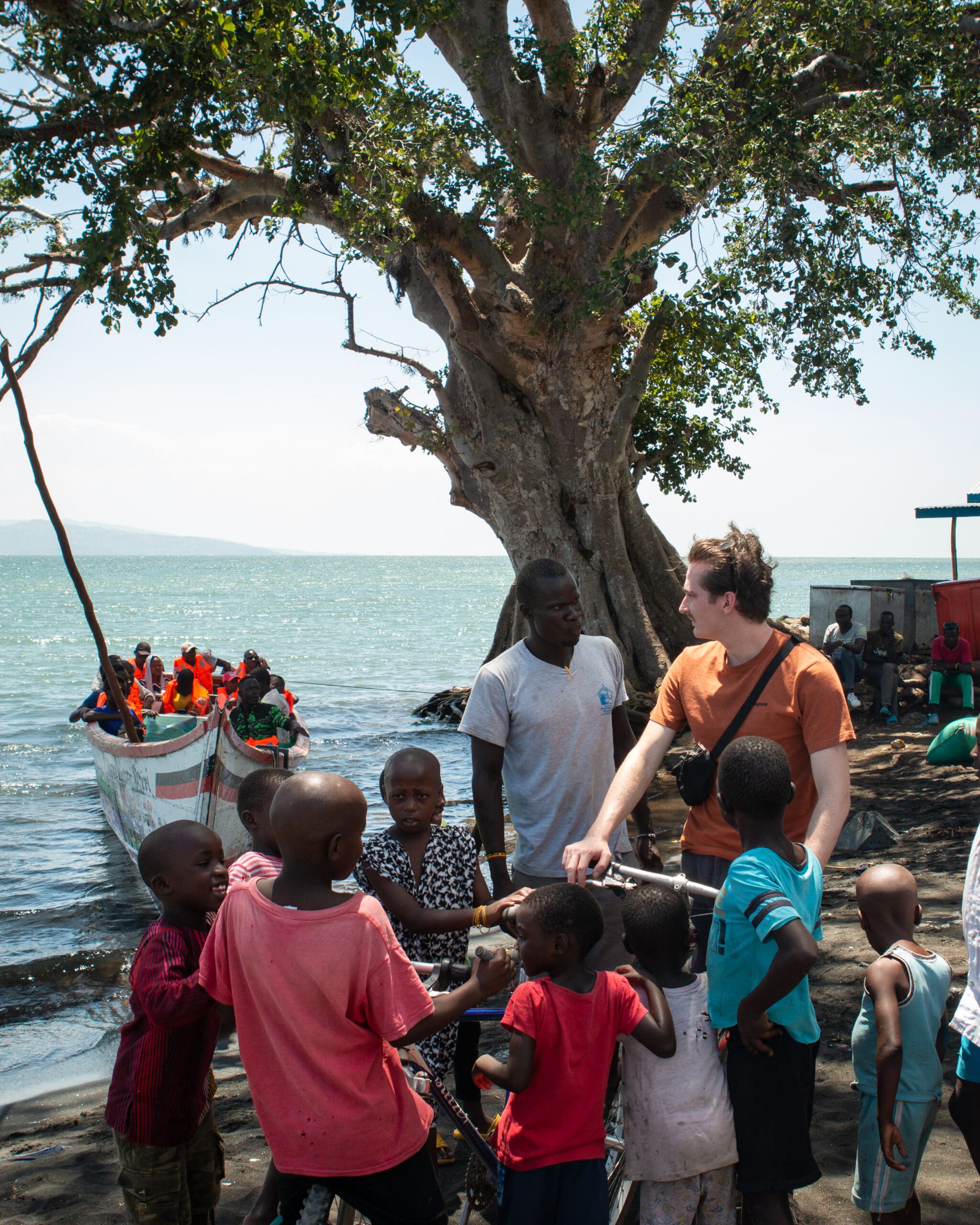 groep kinderen en volwassenen bij het meer van victoria terwijl een boot met mensen aanmeert in kenia