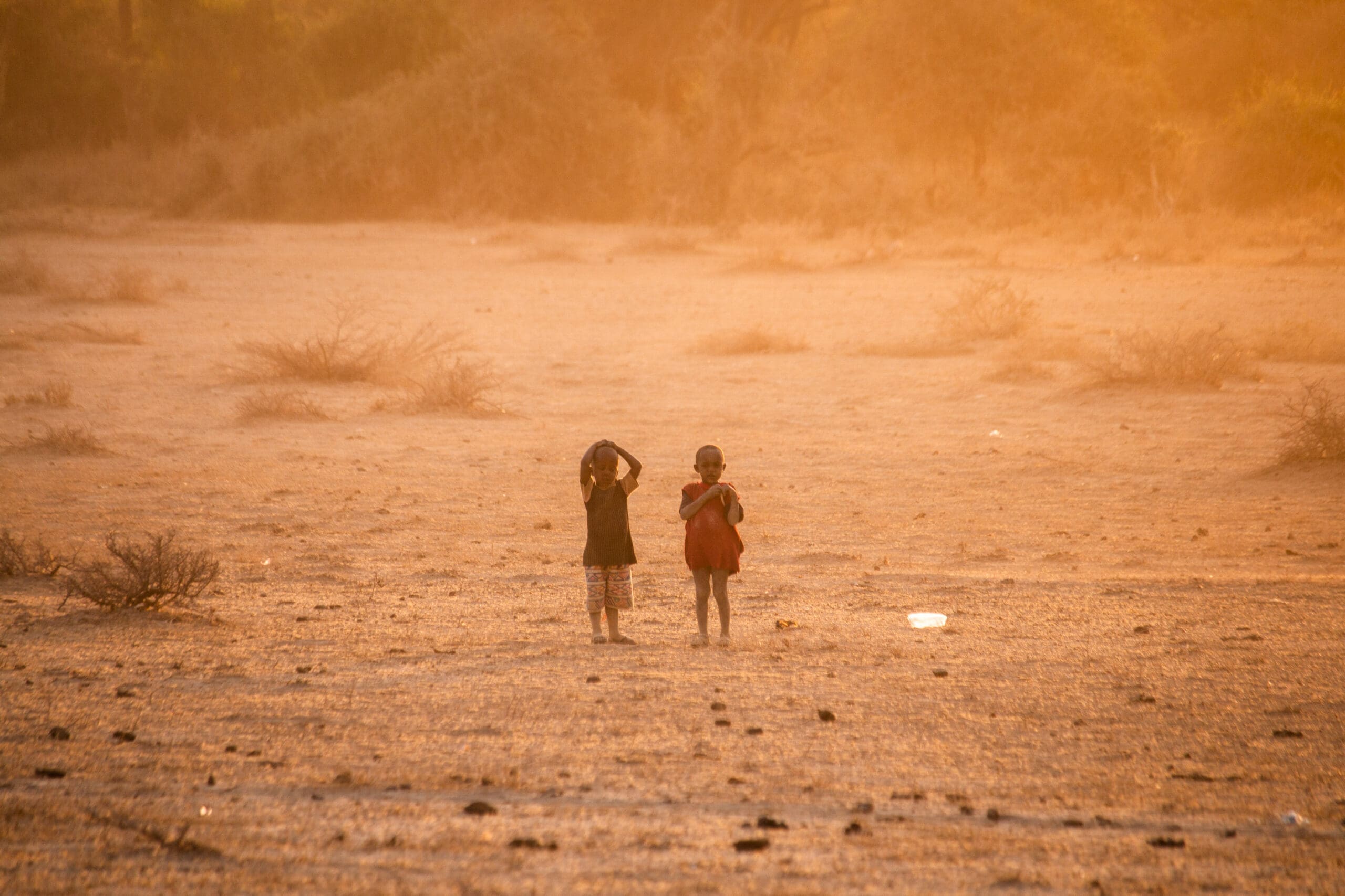 Two young children stand barefoot on a dry, dusty field at sunset, surrounded by sparse bushes and warm golden light, with their backs facing the camera.