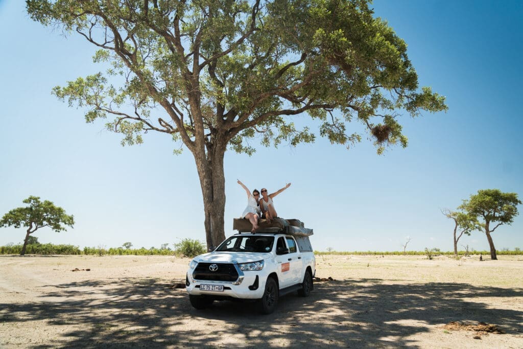 Twee mensen zitten bovenop een witte SUV met kampeerspullen, glimlachen en steken hun armen omhoog onder een grote boom in zonnig Botswana. De helderblauwe lucht en het weinige groen geven de geest van avontuur weer in het open landschap van Afrika.