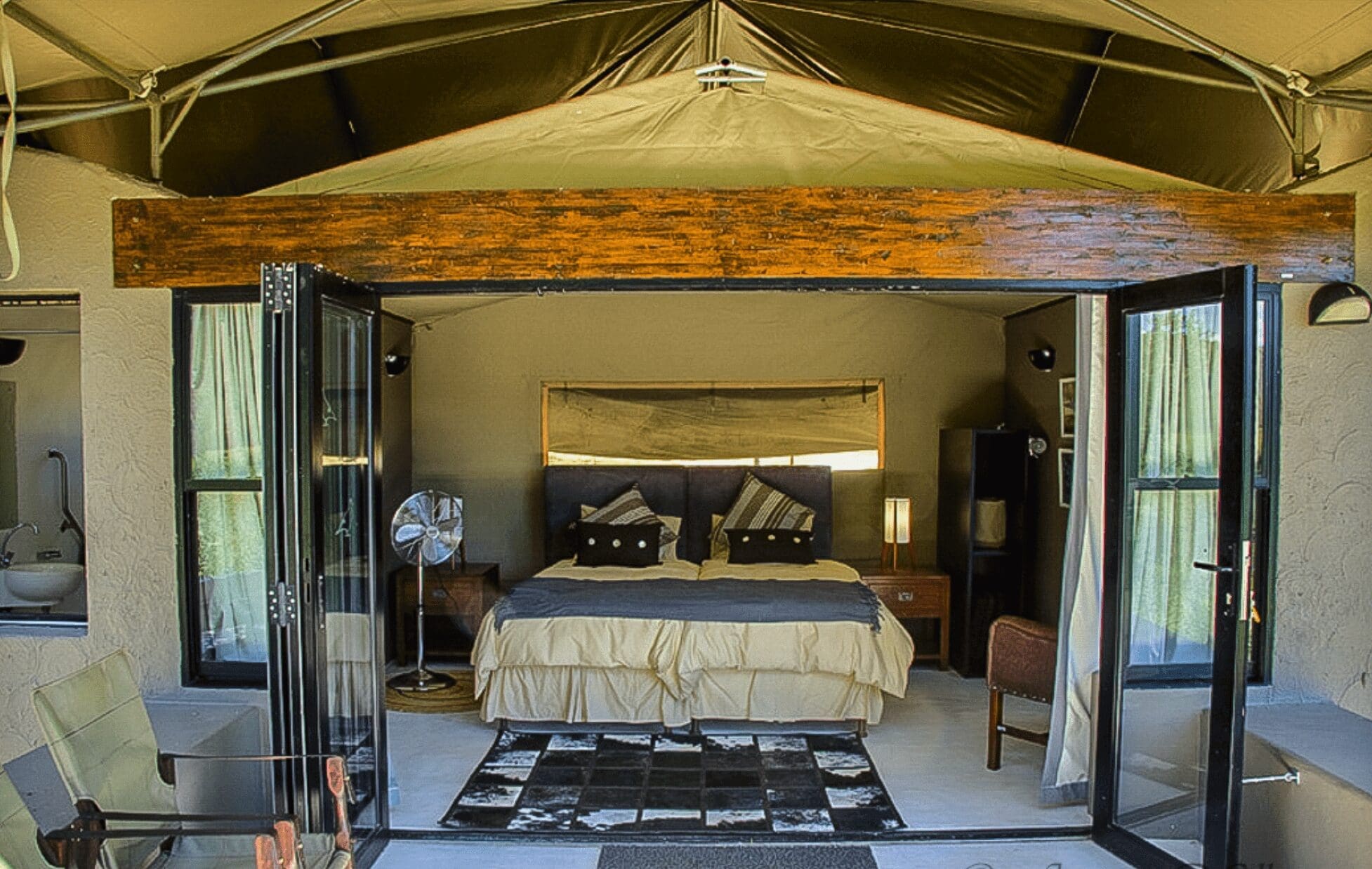 Een ruime, moderne tentkamer in Zuid Afrika met een open ingang toont een netjes opgemaakt bed, bijzettafeltjes met lampen, een ventilator, stoel en gedessineerd tapijt. Natuurlijk licht vult het gezellige interieur - perfect voor reizigers die op zoek zijn naar comfort en avontuur.
