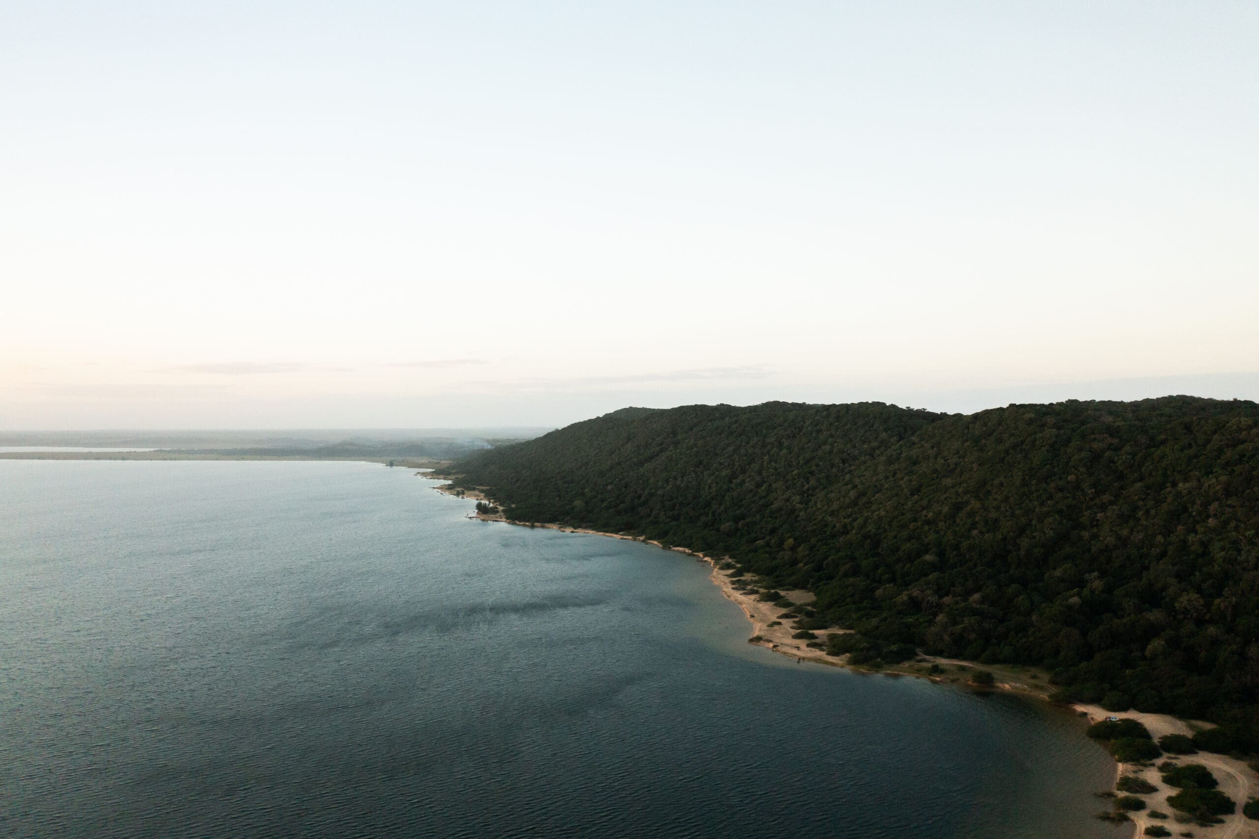 Luchtfoto van een kalme kustlijn met zachte golven, zandstrand en dichtbegroeide groene beboste heuvels die zich in de verte uitstrekken onder een heldere, bleke hemel.