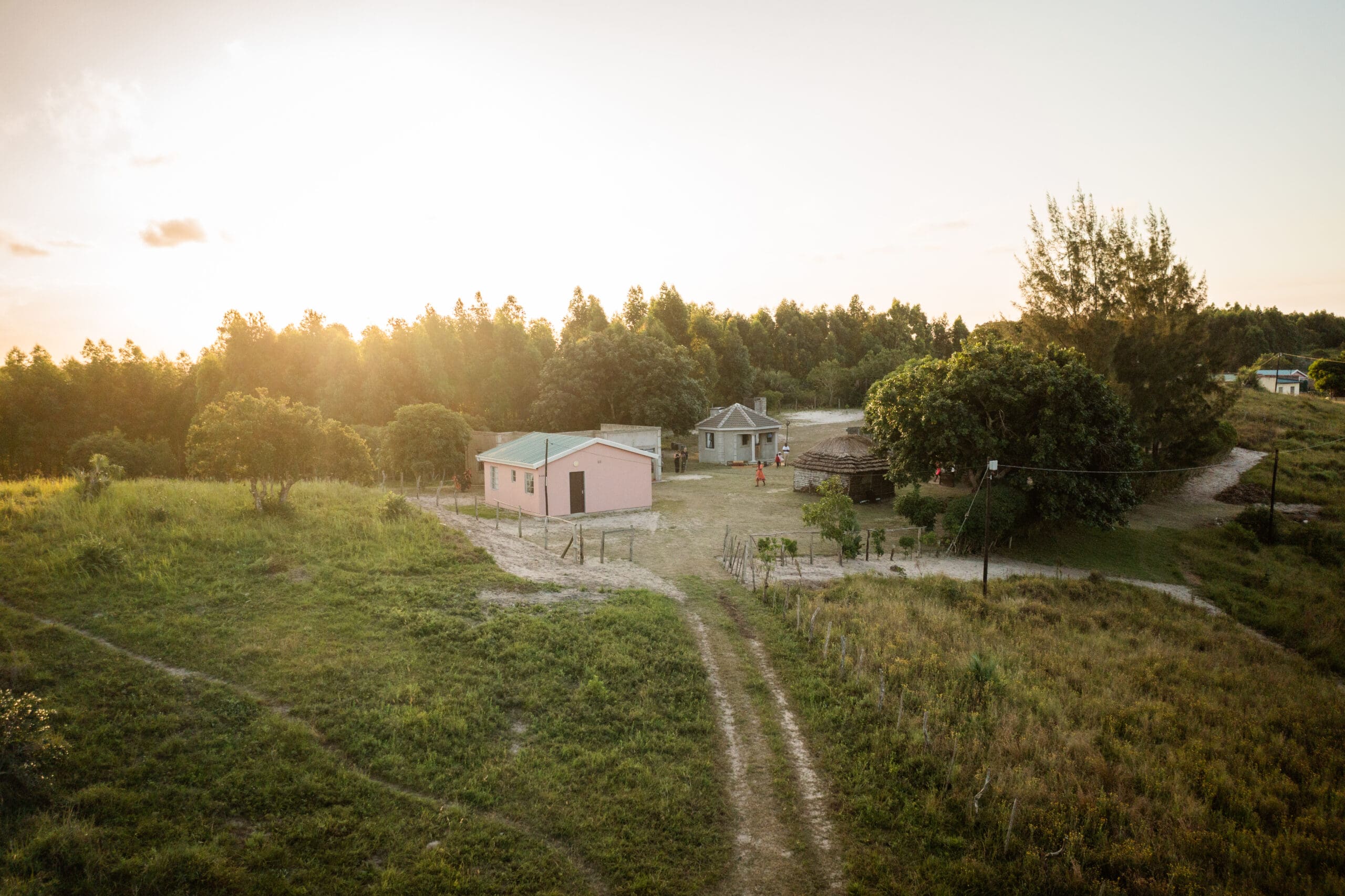 Een landelijk landschap bij zonsondergang toont een roze huis en een ander gebouw omringd door bomen en grasvelden, met onverharde paden die naar de gebouwen leiden. De zon staat laag aan de hemel en werpt een warme gloed.