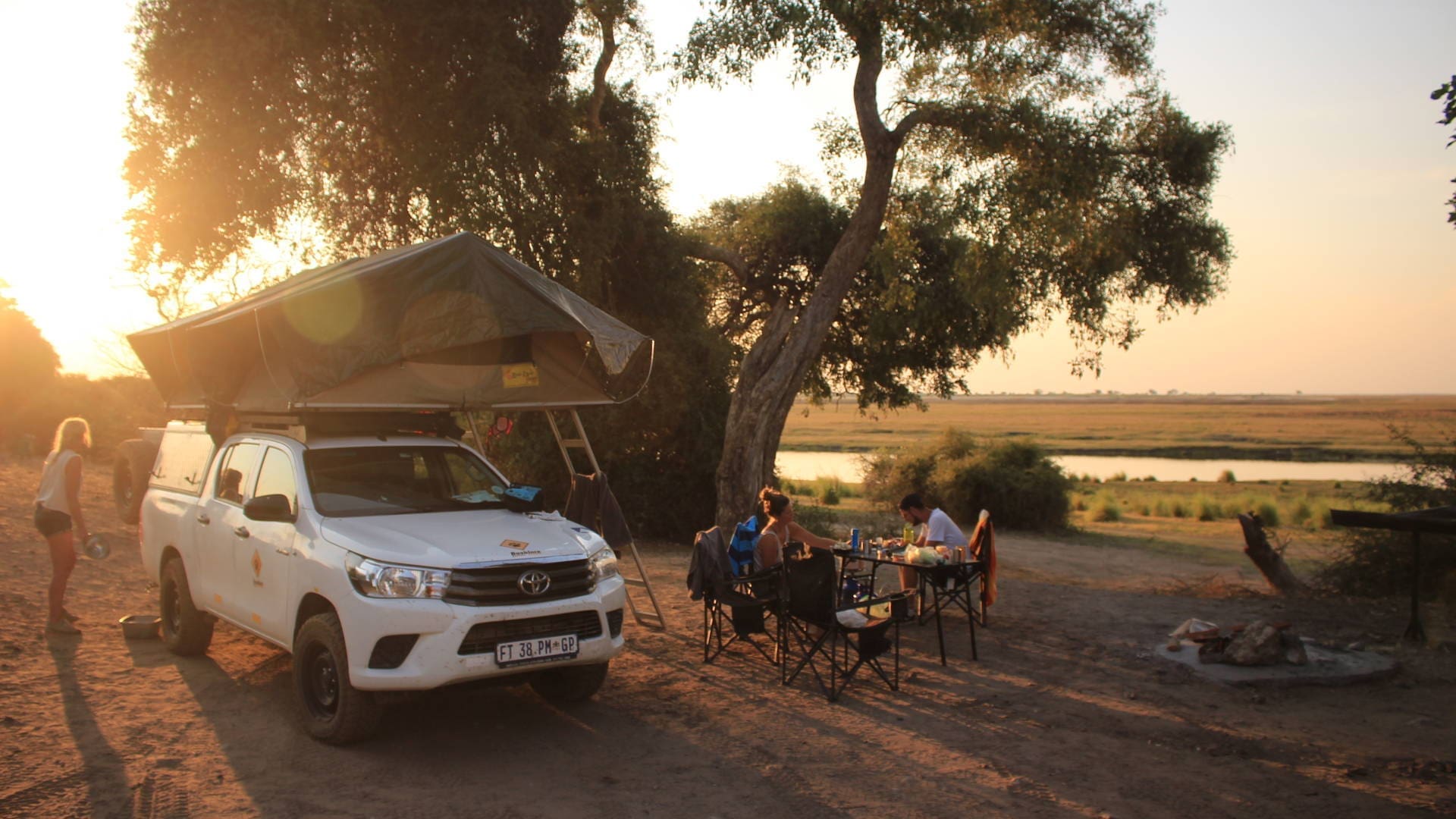 Een witte Toyota truck met een daktent staat geparkeerd op een camping naast een rivier bij zonsondergang. Verschillende mensen zitten aan een tafel te eten of te praten, omringd door bomen en kampeerspullen.