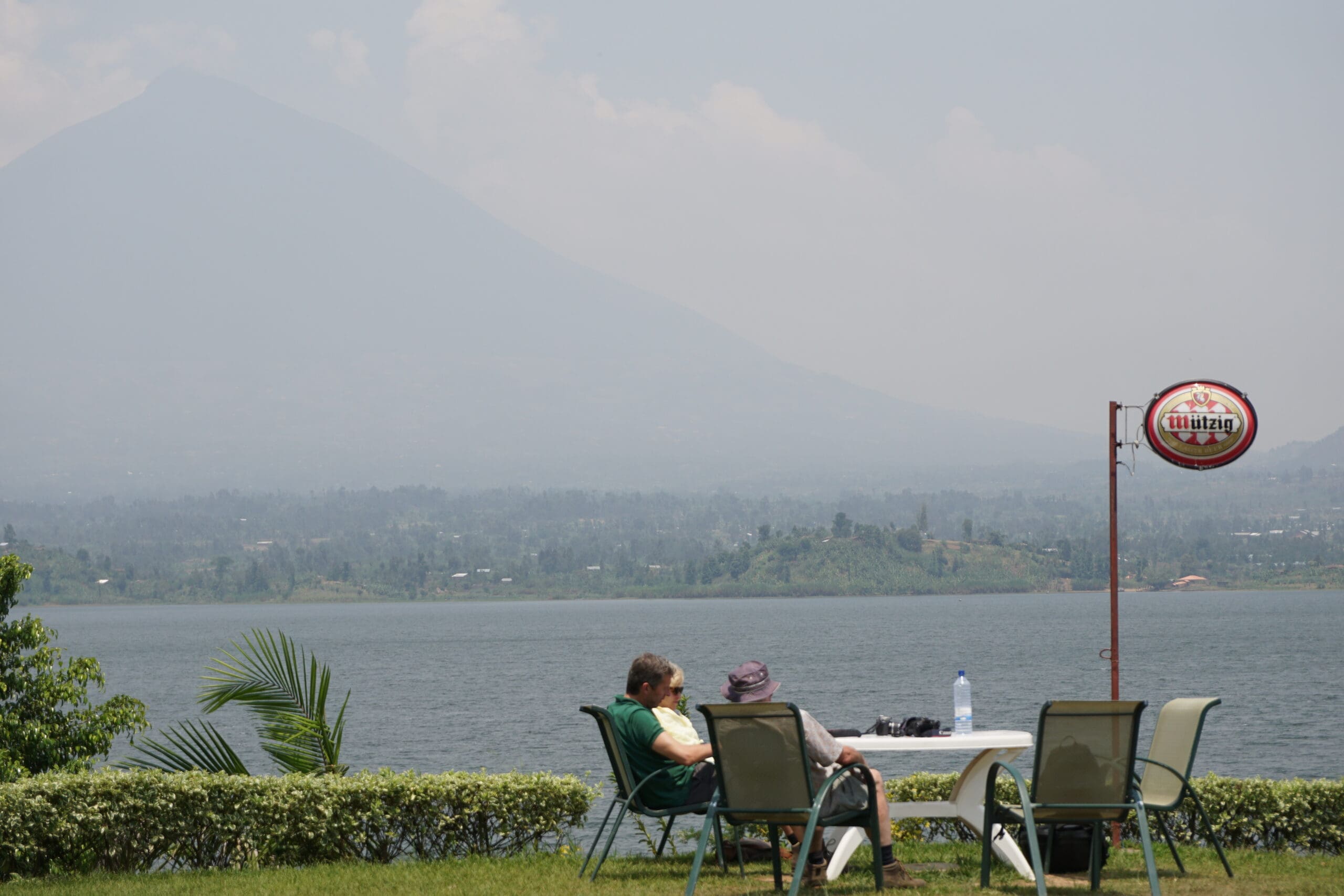 Twee mensen zitten op stoelen aan een tafel met uitzicht op een meer, met een berg zichtbaar in de wazige achtergrond. Er zijn lege stoelen, een fles water op tafel en een wegwijzer in de buurt. Het tafereel ziet er vredig en ontspannen uit.
