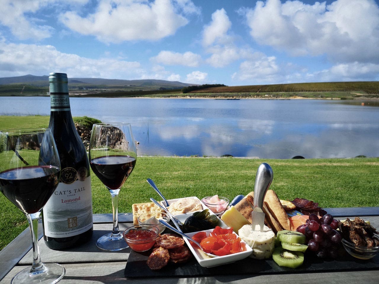 Een picknick bij een meer met twee glazen rode wijn, een fles Cat's Tail wijn en een schotel met kazen, crackers, vleeswaren, fruit en beleg op een houten tafel, met velden en heuvels op de achtergrond.