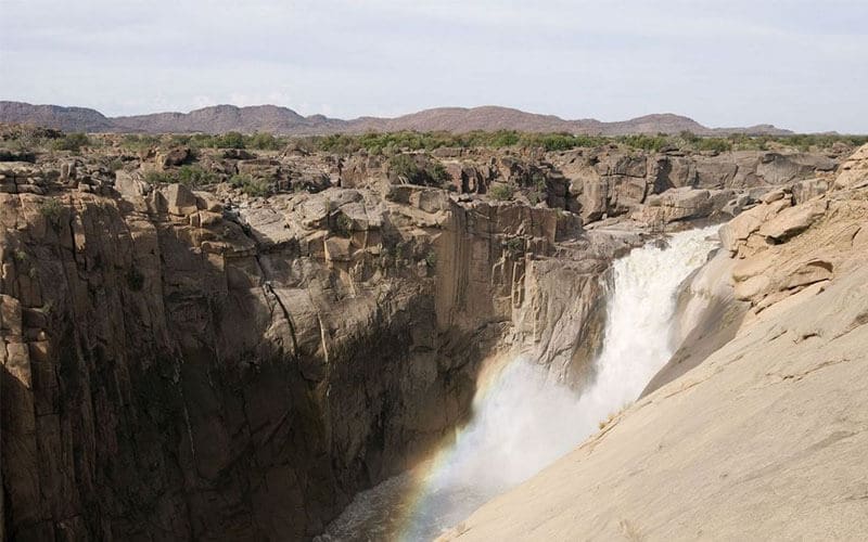A powerful waterfall cascades down a rocky cliff, creating mist and a faint rainbow at the bottom, with rugged terrain and distant hills in the background under a cloudy sky.