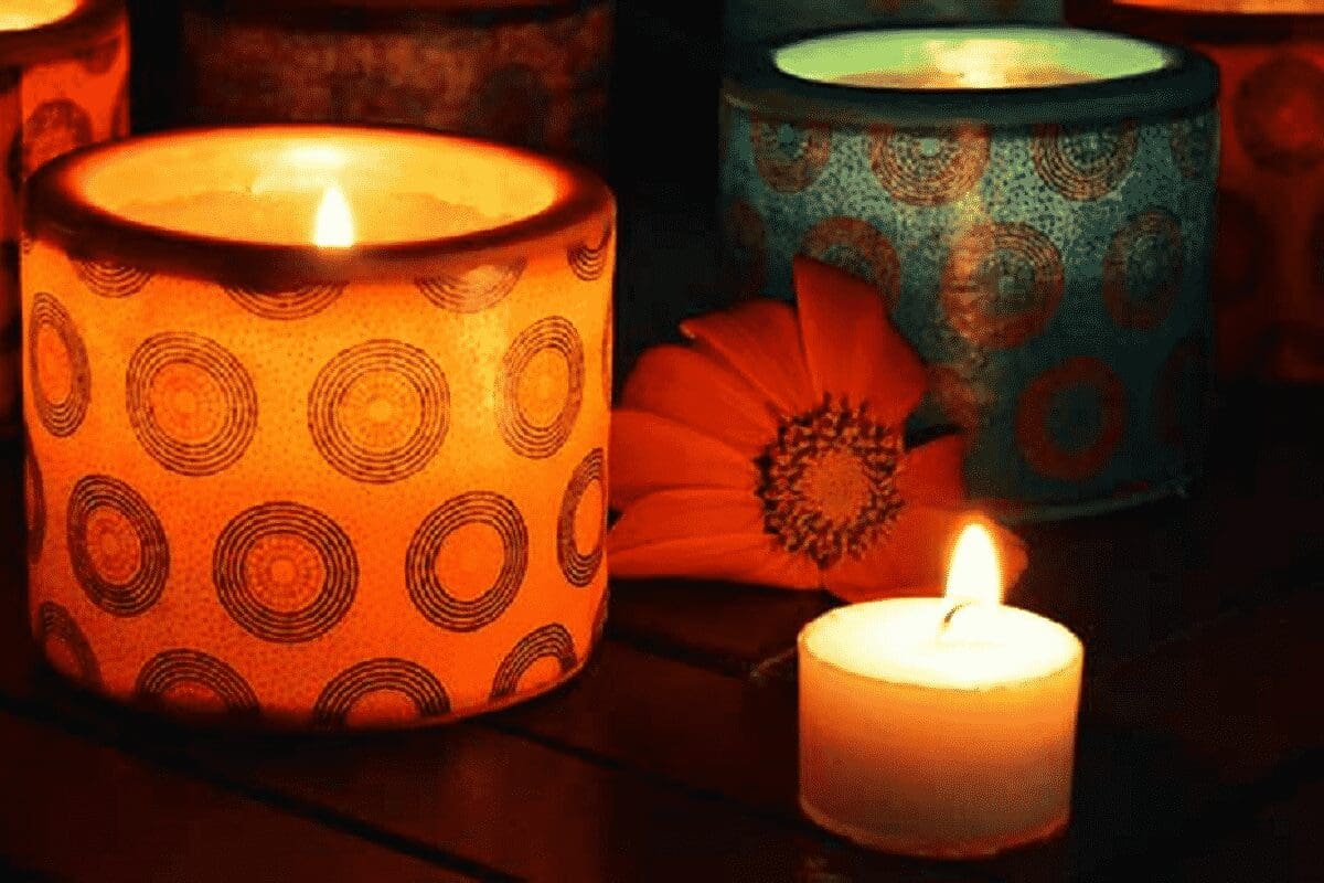Mehrere brennende Kerzen, darunter eine große orangefarbene Kerze in einem gemusterten Halter mit kreisförmigen Mustern und eine kleine Teelichtkerze, stehen neben einer orangefarbenen Blume auf einer dunklen Holzfläche.