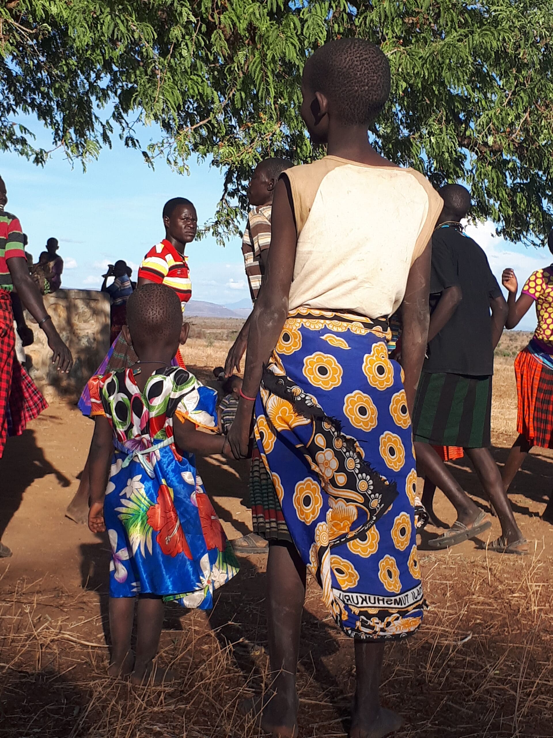 Een vrouw en kind, beiden in kleurrijke kleding met patronen, houden elkaars hand vast in een levendige groep buiten onder een boom in Oeganda, Afrika. Dit is een glimp van de levendige sfeer die je kunt ervaren als je door deze prachtige regio reist.