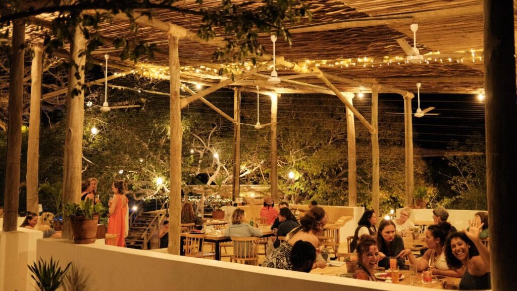 An outdoor restaurant at night with string lights overhead, wooden beams, and people dining at tables. The atmosphere is warm and lively, evoking the spirit of travelling in Kenya, with trees visible in the background.