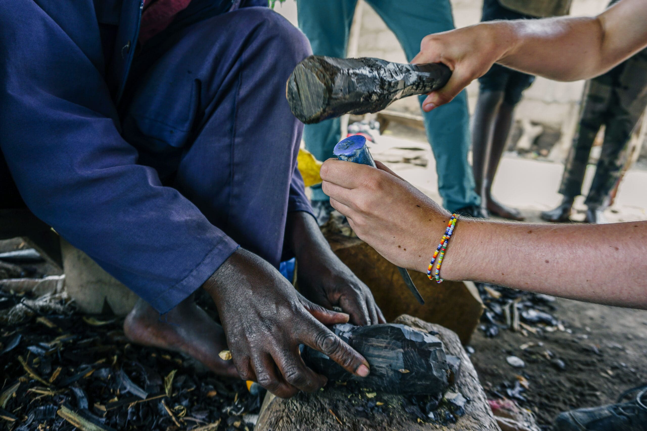 Two people shape a piece of black stone; one holds it steady while the other strikes with a hammer. The image shows close-up hands, one wearing a colorful beaded bracelet, surrounded by stone debris.