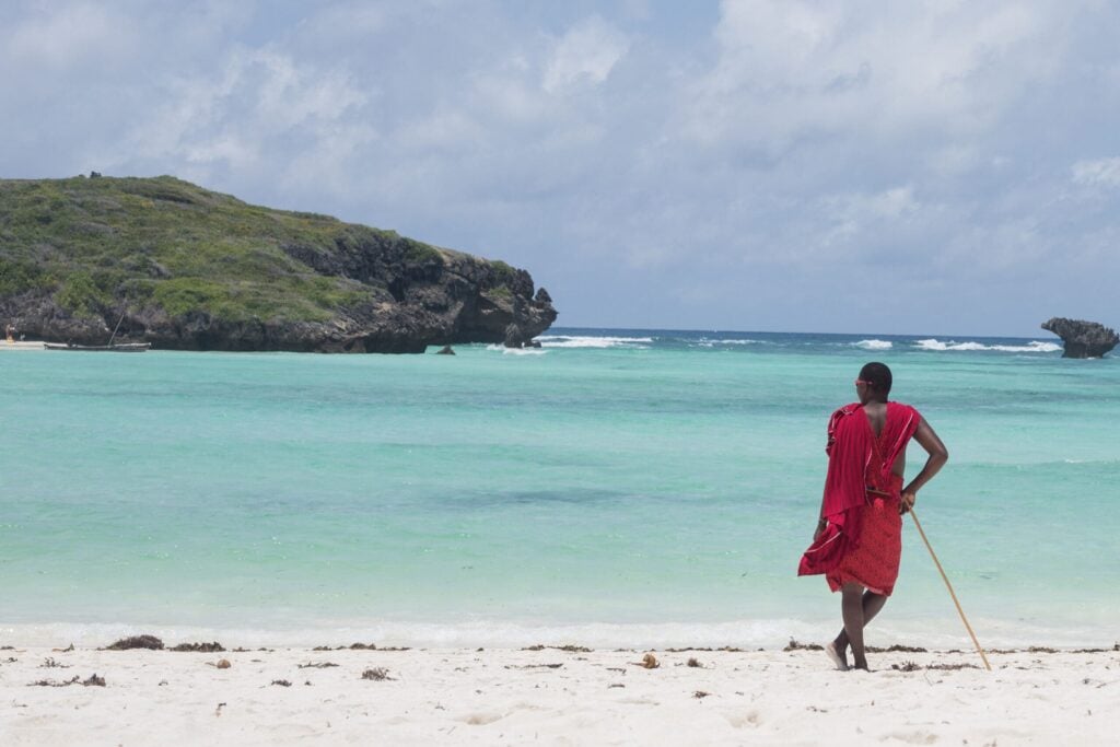 masai in rode shuka kijkt uit over turquoise oceaan bij rotsachtige kust in Kenia