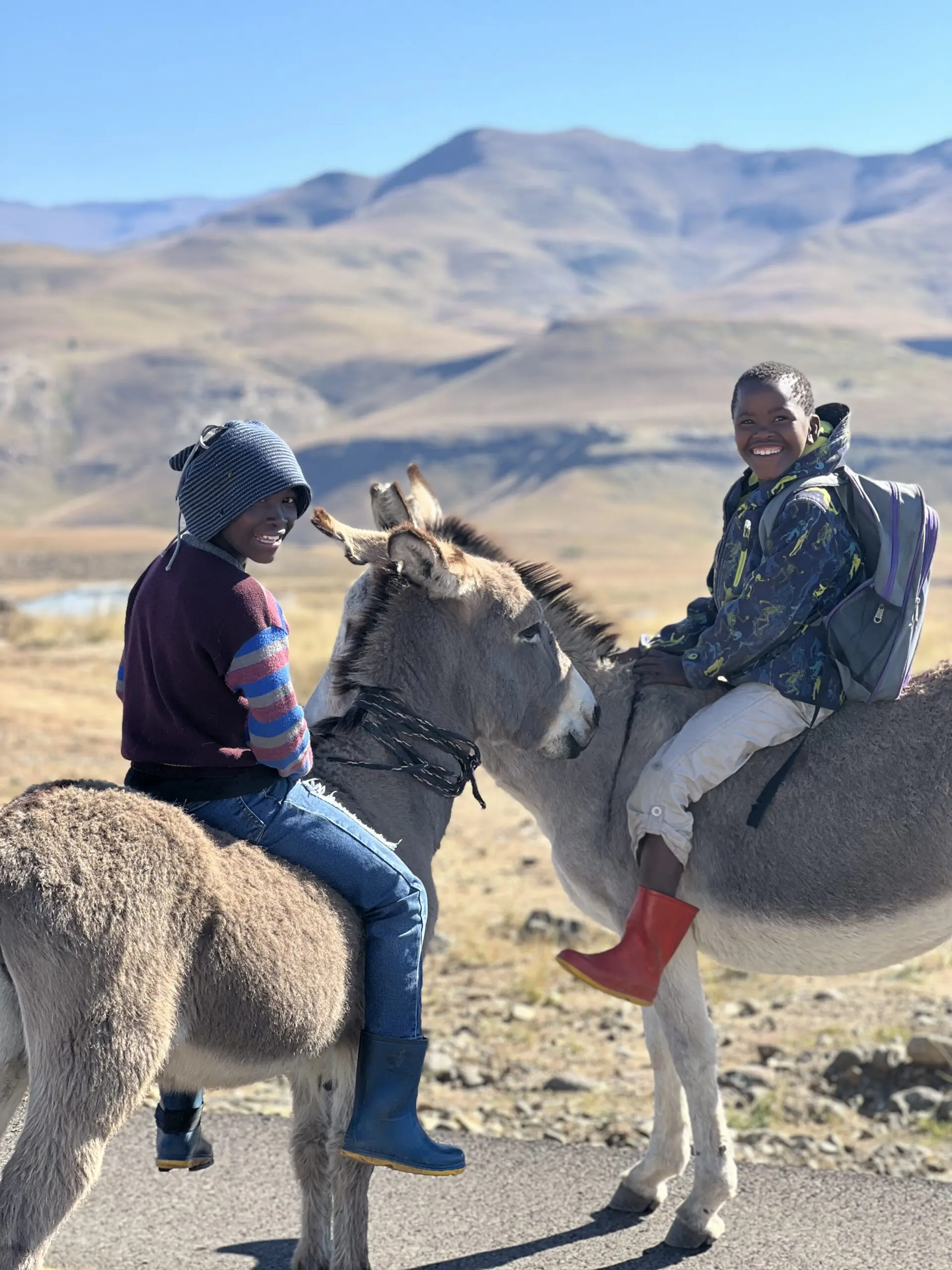 Two smiling children sit on donkeys in a hilly, sunlit landscape. One child wears a hat, striped sweater, and blue boots, the other wears a camouflage jacket, backpack, and red boots. Mountains rise in the background.