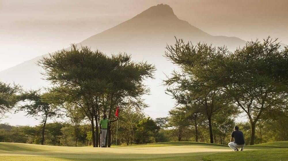 Twee mensen op een golfbaan in Tanzania, omringd door bomen, met de een aan het putten en de ander gehurkt in de buurt. Een grote berg rijst op in de achtergrond onder een wazige, gouden Afrika-hemel die perfect is voor liefhebbers van reizen.