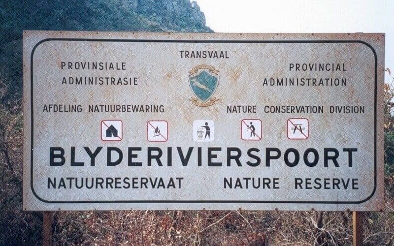 Ein verwittertes Schild für das Blyde River Canyon Nature Reserve in Afrikaans und Englisch mit Vorschriften zum Verbot von Feuer, Zelten, Haustieren und Müll, vor einem bergigen Hintergrund.