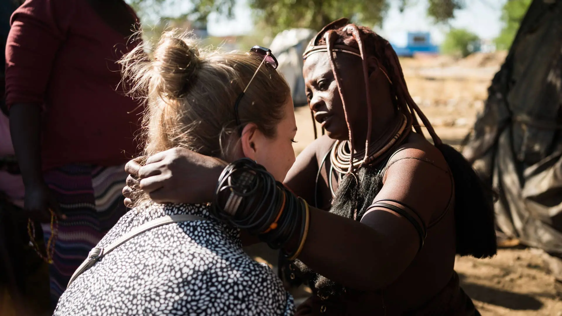 A Himba woman with traditional jewelry and hair touches the shoulders of a woman with light hair in a bun, who is sitting and facing her, outdoors in sunlight.