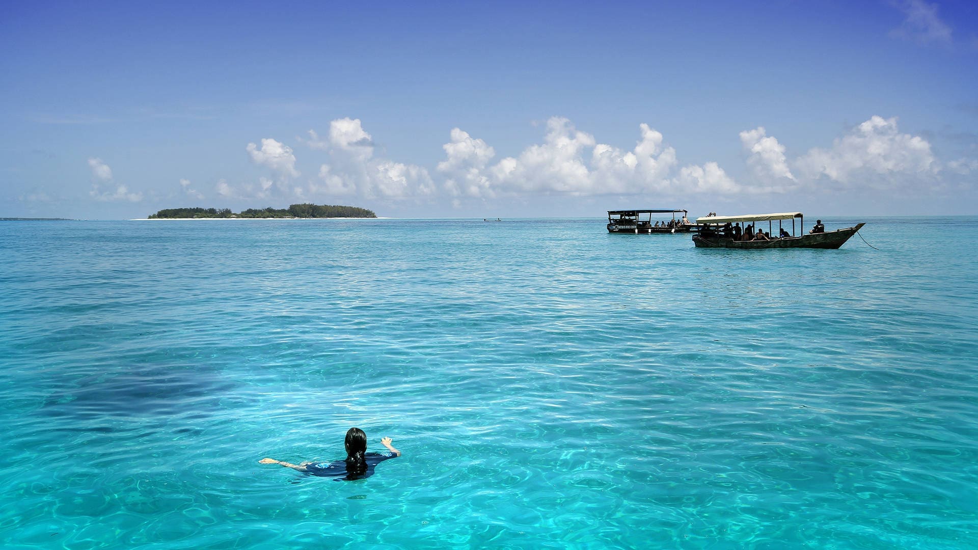 A person swims alone in clear turquoise water near a distant island, with two boats and a blue sky dotted with white clouds—capturing the essence of travelling in Tanzania, Africa.