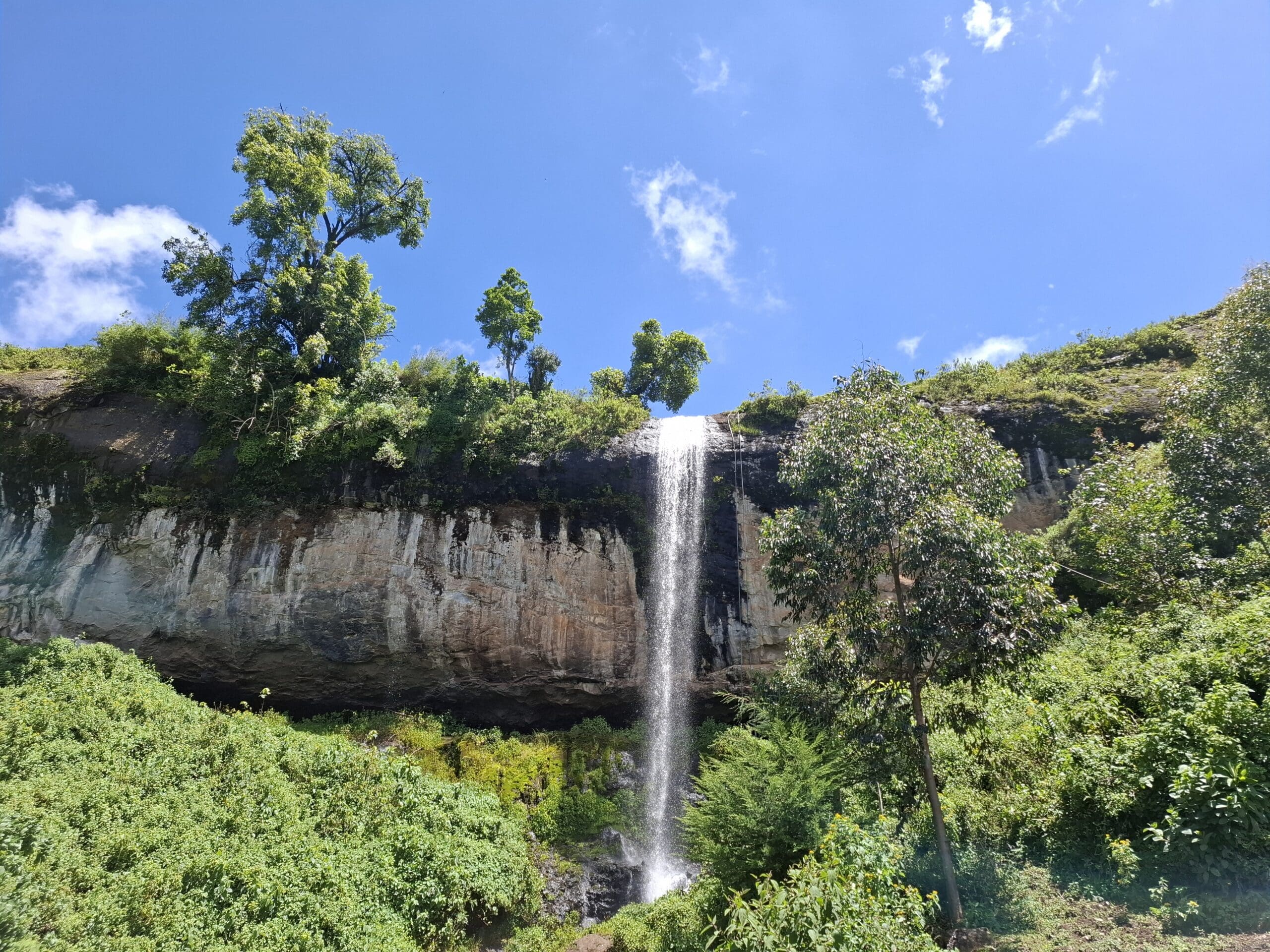 Een hoge, smalle waterval stort zich uit over een rotsachtige klif omgeven door weelderige groene vegetatie onder een helderblauwe hemel met een paar wolken - een verborgen paradijs in Oeganda, Afrika, dat wacht op avontuurlijke reizigers om de schoonheid ervan te ontdekken.