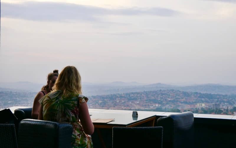Twee vrouwen zitten aan een buitentafel met menu's en kijken uit over een stadslandschap en heuvels in de verte onder een bewolkte hemel - een uitnodigend tafereel dat doet dromen van reizen door Afrika of de prachtige landschappen van Oeganda.