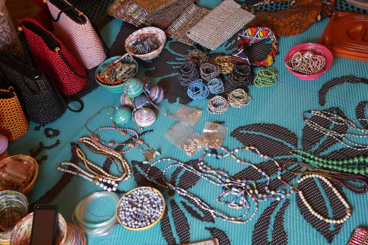 Een verscheidenheid aan handgemaakte kralen sieraden, kleurrijke portemonnees en ambachtelijke benodigdheden uit Afrika zijn verspreid over een helder blauw patroon stof, met kettingen, armbanden en kleine tassen in verschillende kleuren en stijlen.