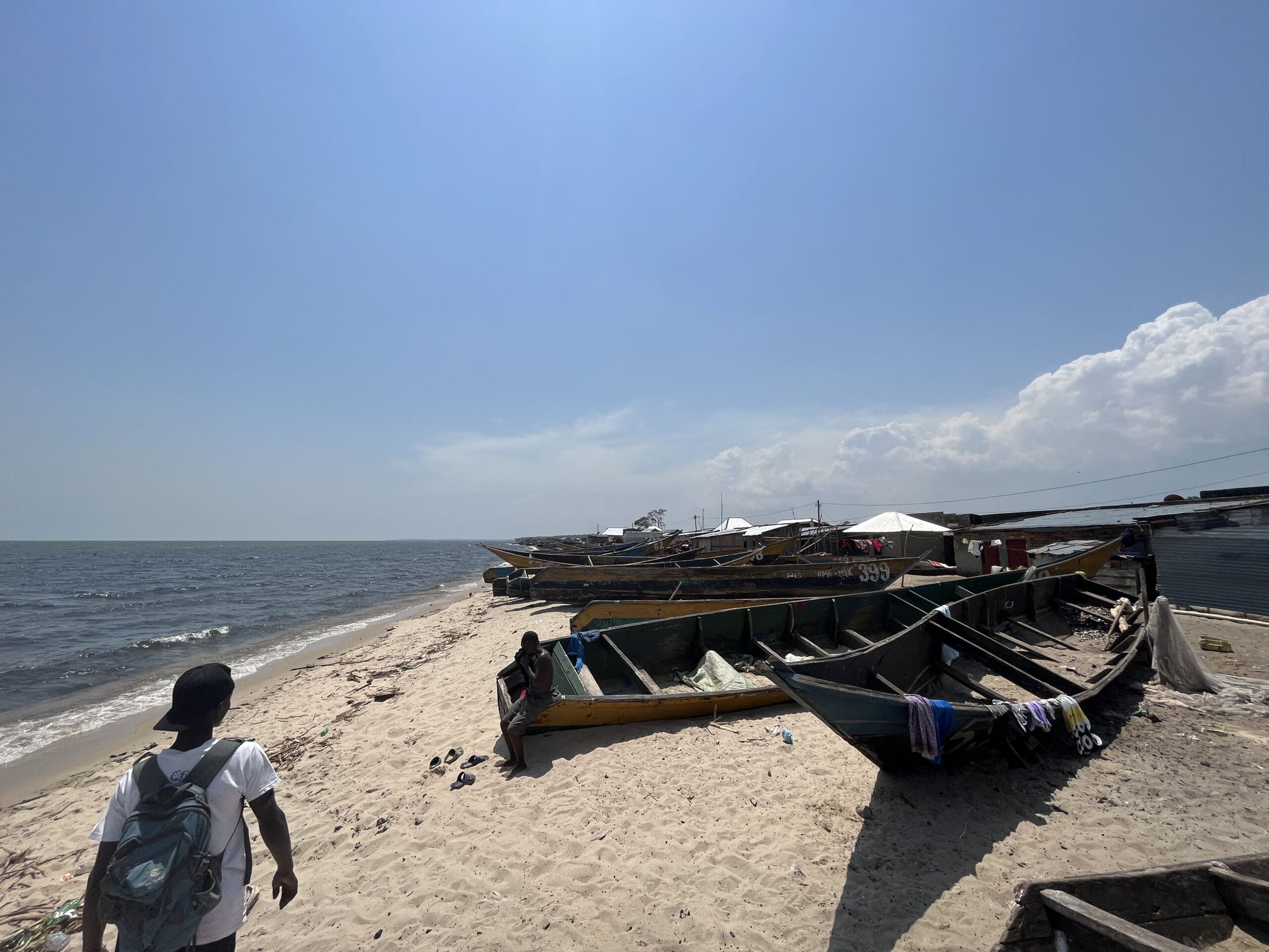 Een zandstrand in Afrika met houten vissersboten langs de kust. Een reiziger met een rugzak loopt naar de boten toe, terwijl een andere persoon in één van de boten zit. De lucht is meestal helder met een paar wolken.