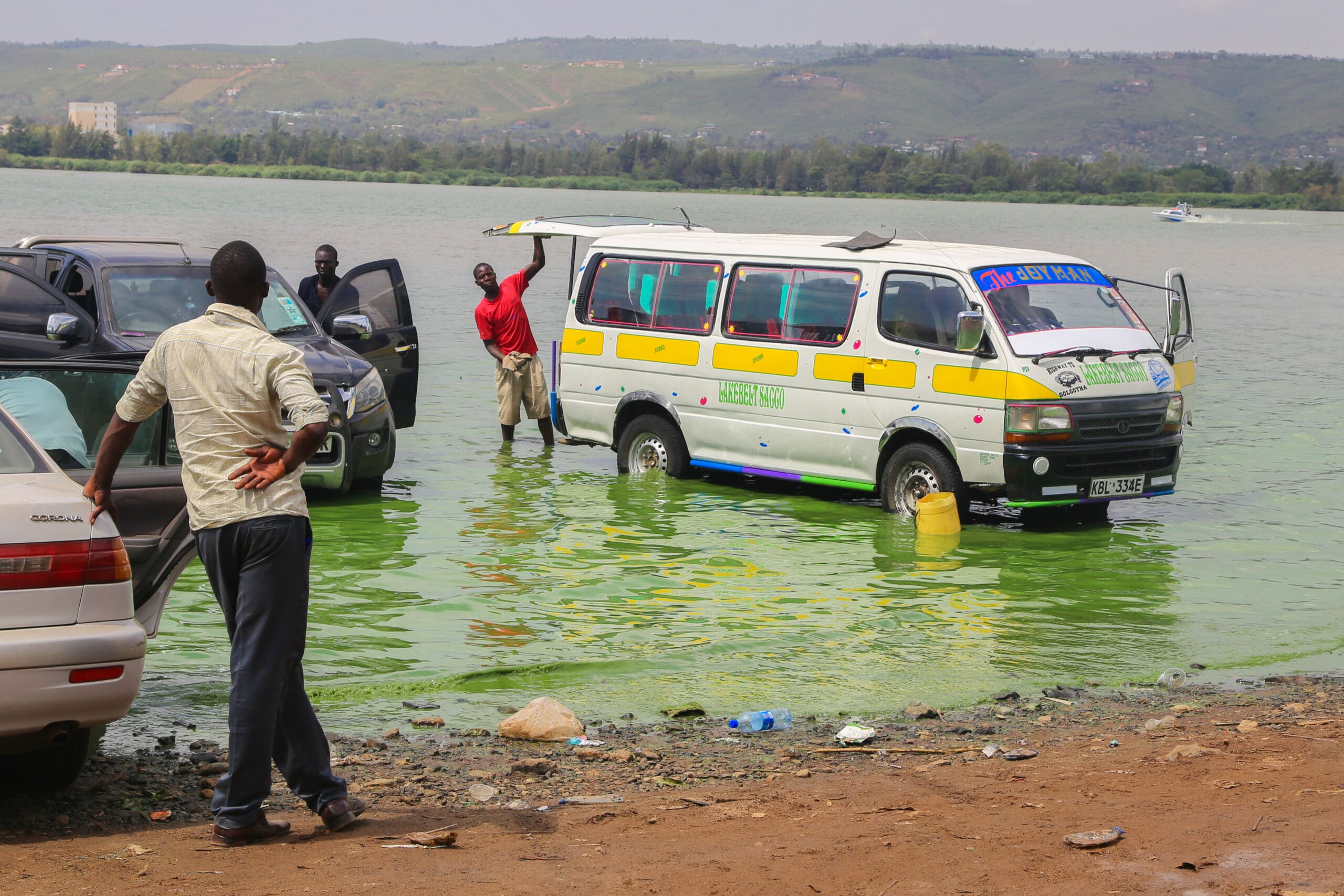 Een man staat aan de oever toe te kijken hoe een minibus en een aantal auto's gedeeltelijk onder water worden gezet in een meer. Een andere man opent de deur van de minibus terwijl hij in het water staat. Op de achtergrond zijn groene heuvels zichtbaar.