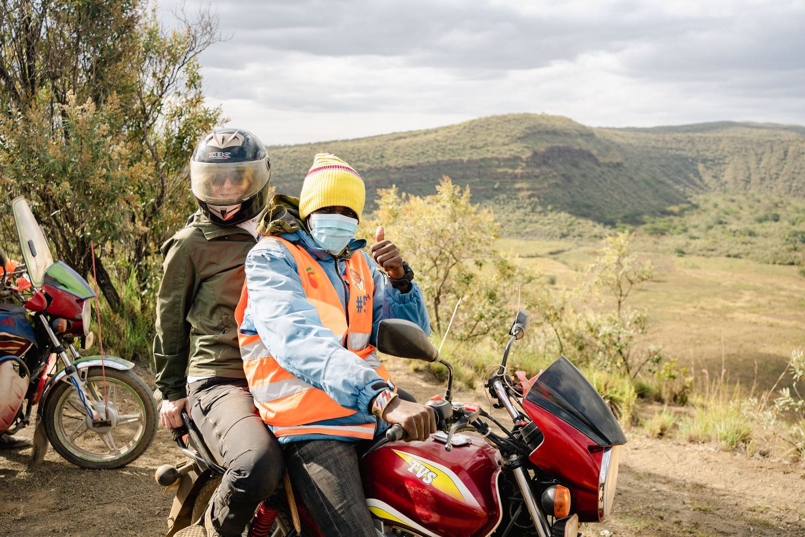 reiziger met helm achterop een boda boda motor met gids in fel oranje hesje in heuvelachtig landschap van kenia