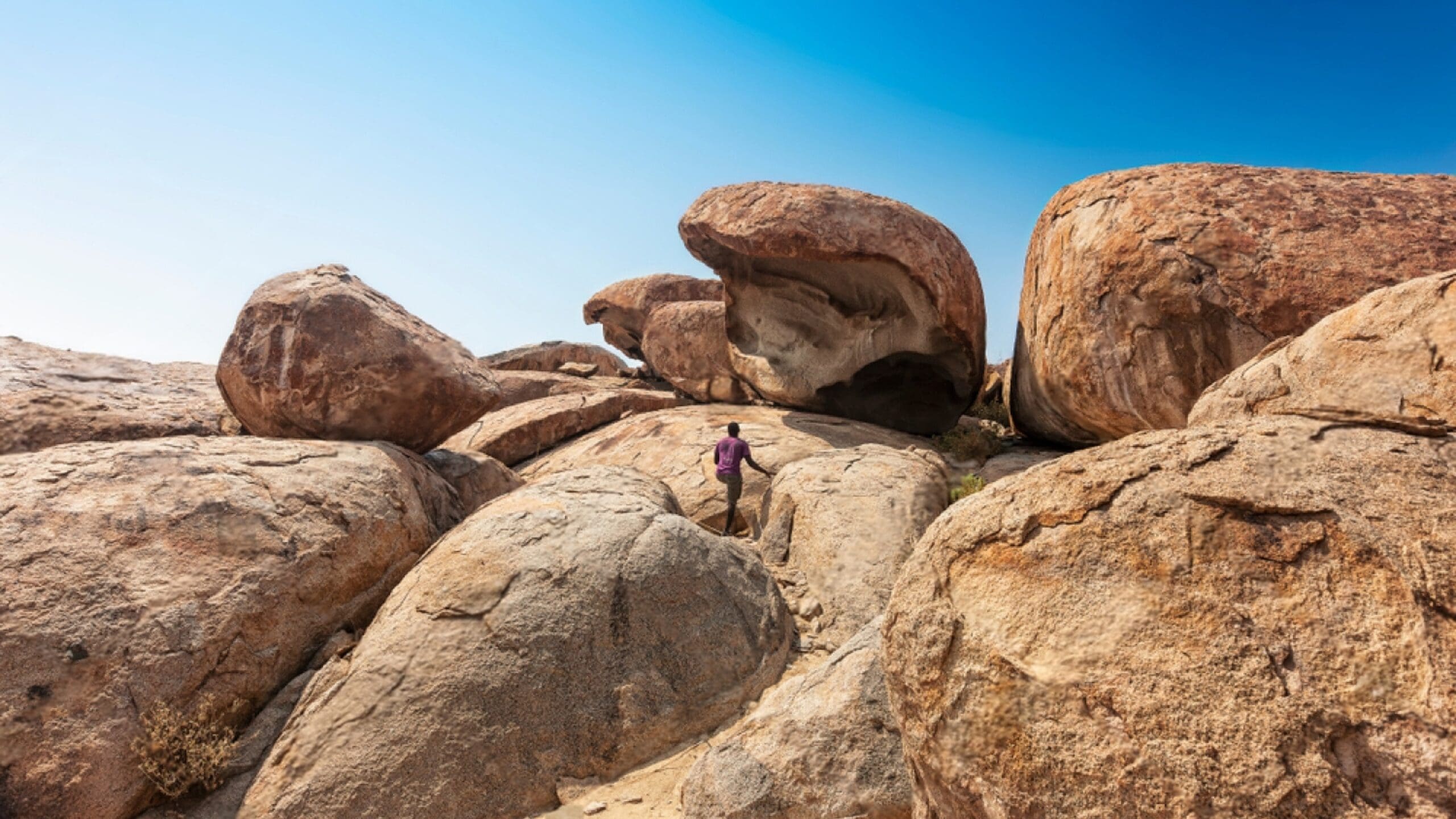 Een reiziger met een paars shirt beklimt grote, ruige, zonovergoten rotsblokken onder een strakblauwe hemel. Enorme rotsformaties domineren het landschap en creëren een opvallend natuurlijk tafereel dat doet denken aan de wilde schoonheid van Angola.