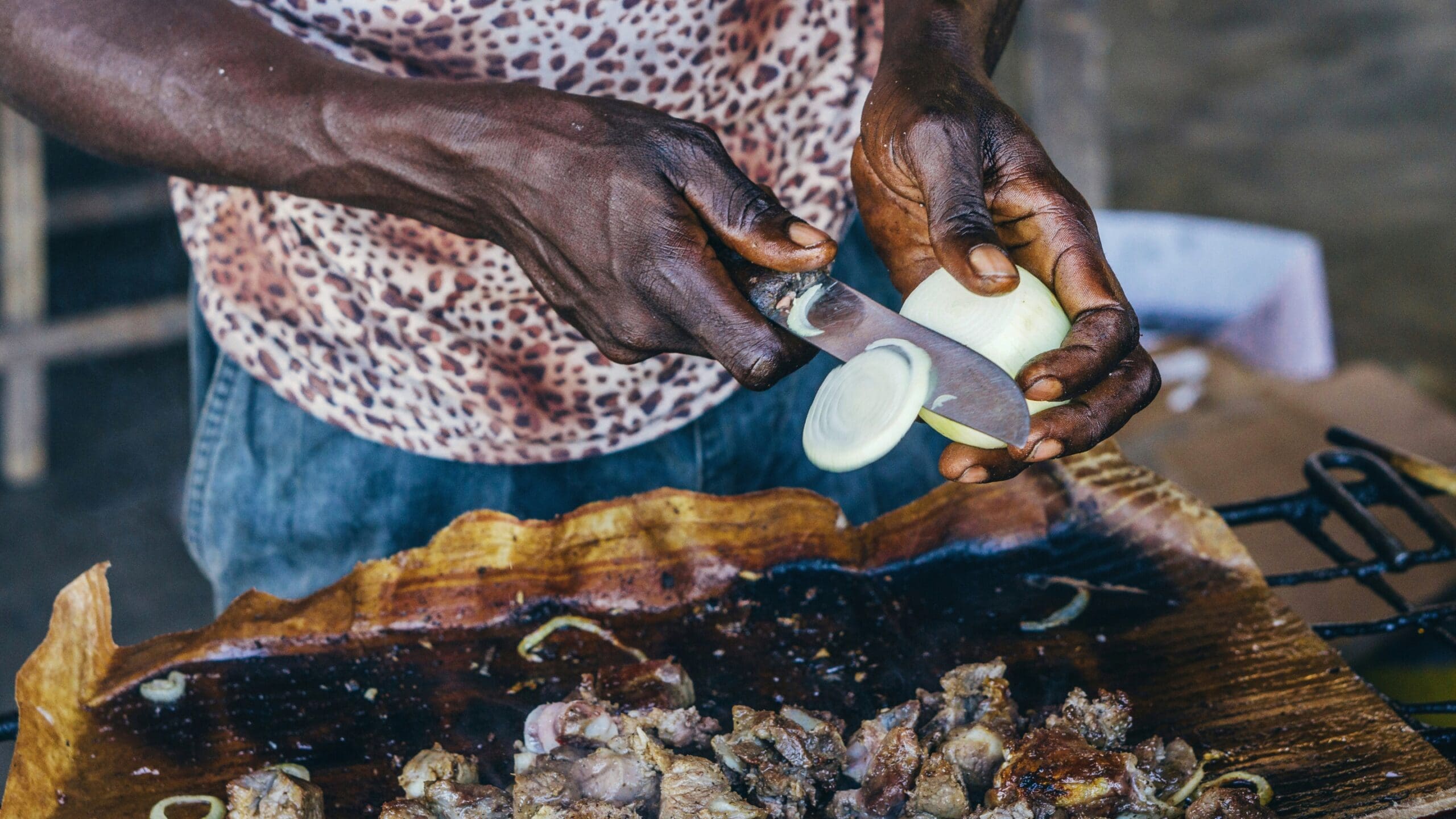 Een persoon in een shirt met luipaardprint snijdt een ui over gegrild vlees en een bruin blad, terwijl hij het eten buiten bereidt - een authentieke reizigerservaring die doet denken aan reizen door Angola.