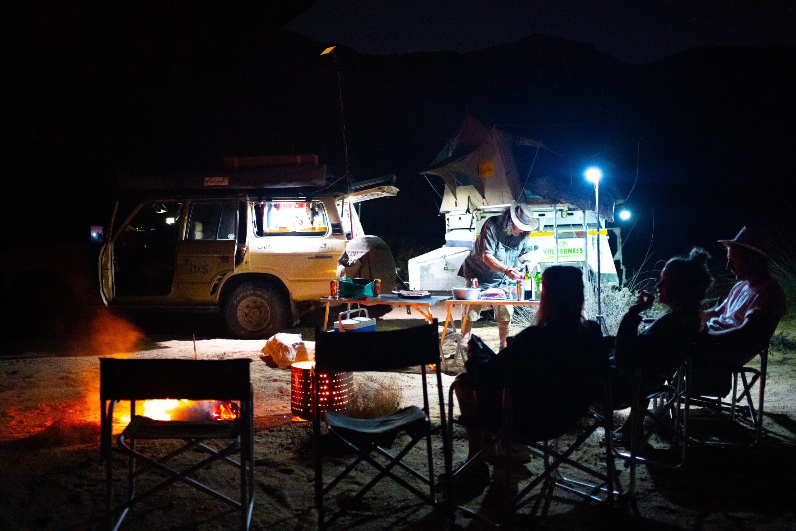 mensen zitten rond een kampvuur in namibië terwijl een man kookt bij een terreinwagen en verlichte aanhanger onder de sterrenhemel