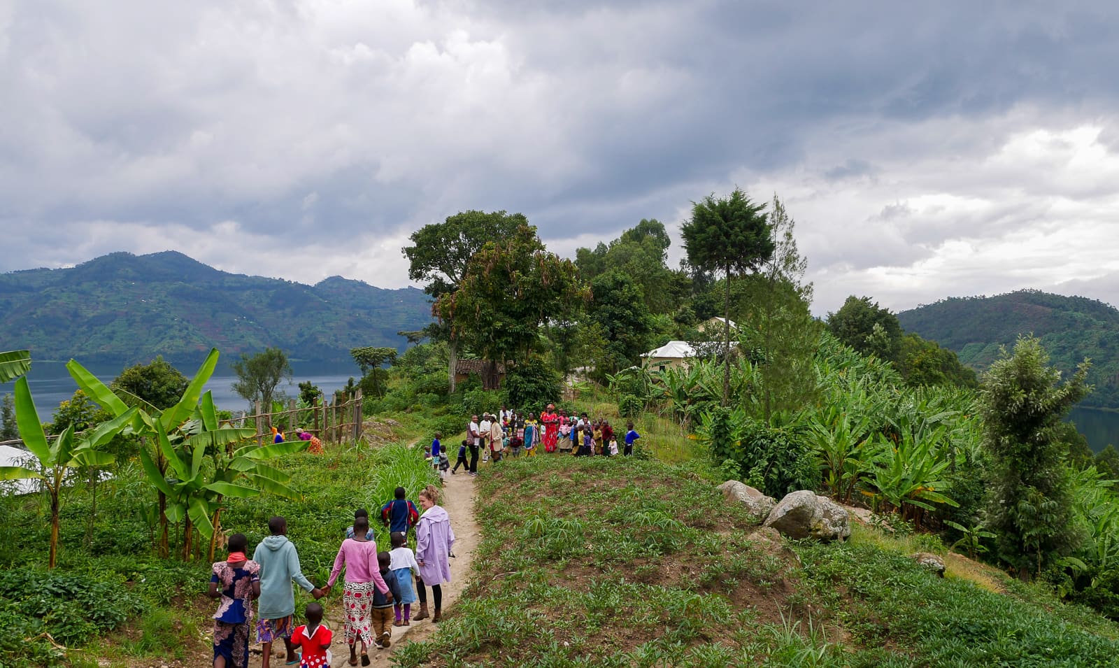 Een groep mensen, waaronder kinderen, loopt over een smal pad op een weelderige Rwandese heuvel bij een meer met bergen op de achtergrond onder een bewolkte Afrikaanse lucht. Bananenplanten en andere vegetatie omzomen het pad.