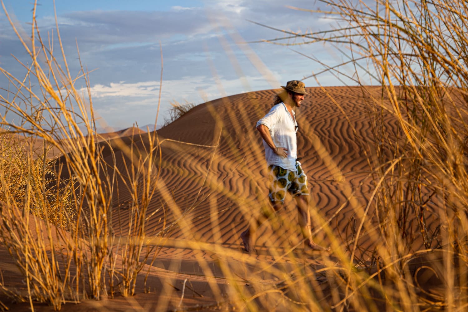 een man wandelt blootsvoets door roodkleurige zandduinen met droge begroeiing in namibië tijdens het gouden uur