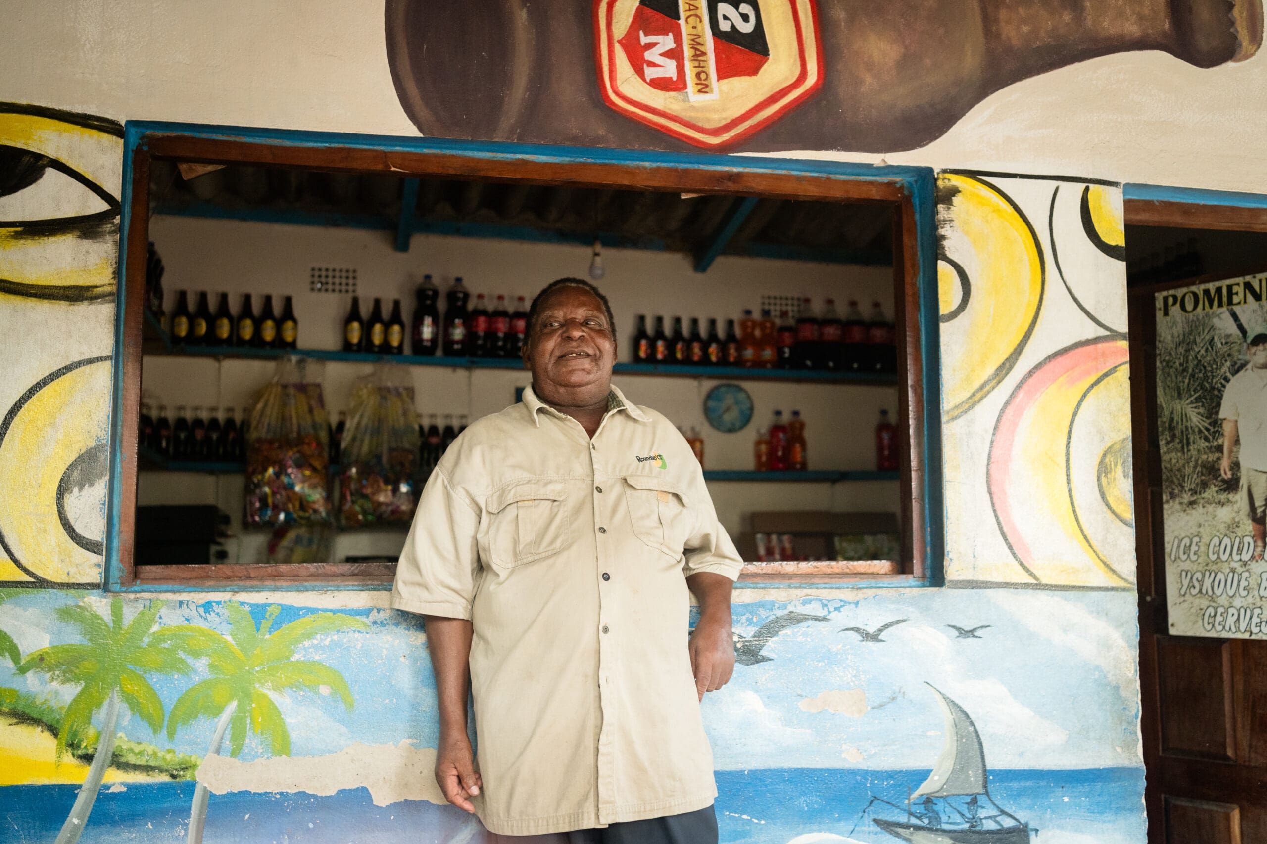 Een man in een beige overhemd staat glimlachend bij een kleurrijke toonbank met een muurschildering op het strand, in een Mozambikaanse winkel vol drankjes en snacks, met helder geschilderde decoraties op de muur achter hem, die de levendige geest van Afrika vastleggen.