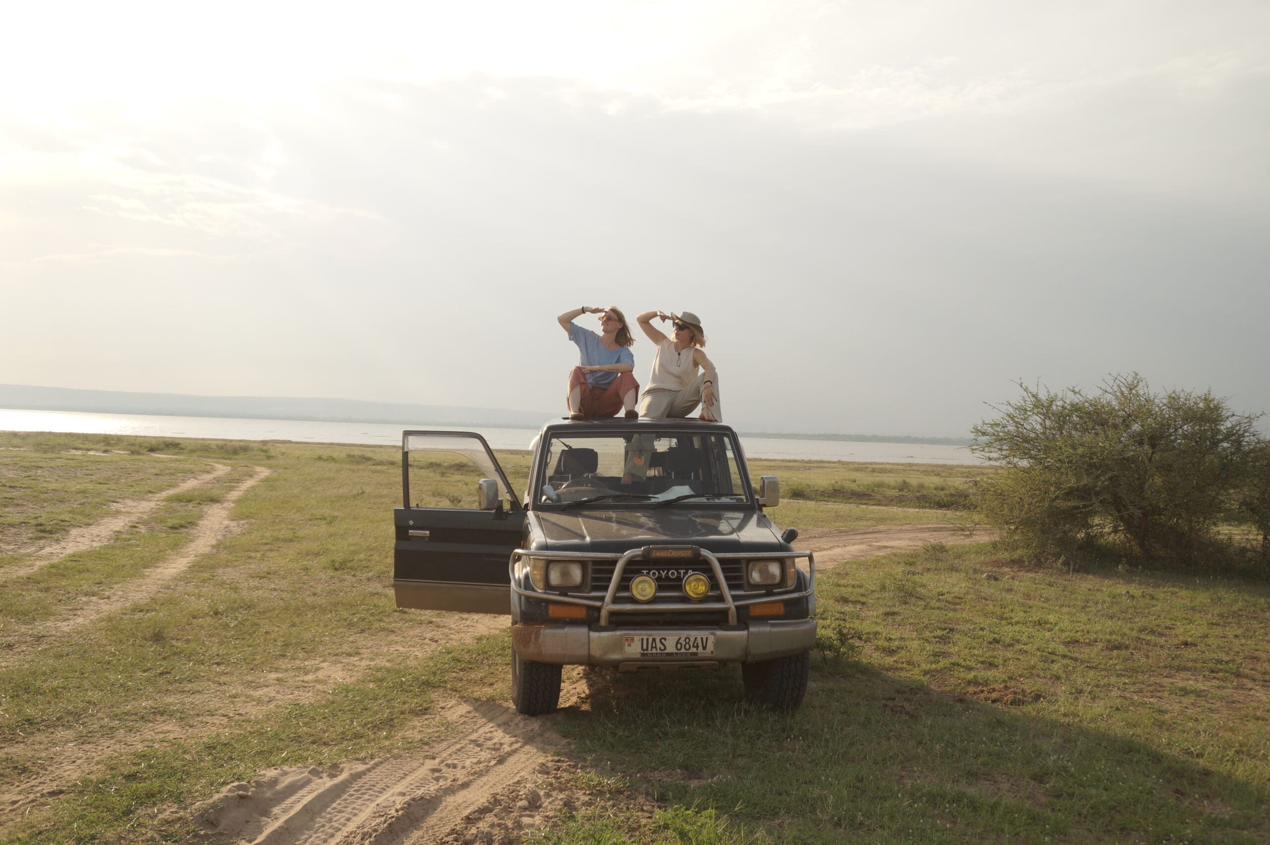 Twee reizigers staan op het dak van een geparkeerde SUV en bedekken hun ogen terwijl ze uitkijken over een open grasvlakte aan het water in Oeganda, Afrika. Een deur hangt open onder de heldere, bewolkte hemel.