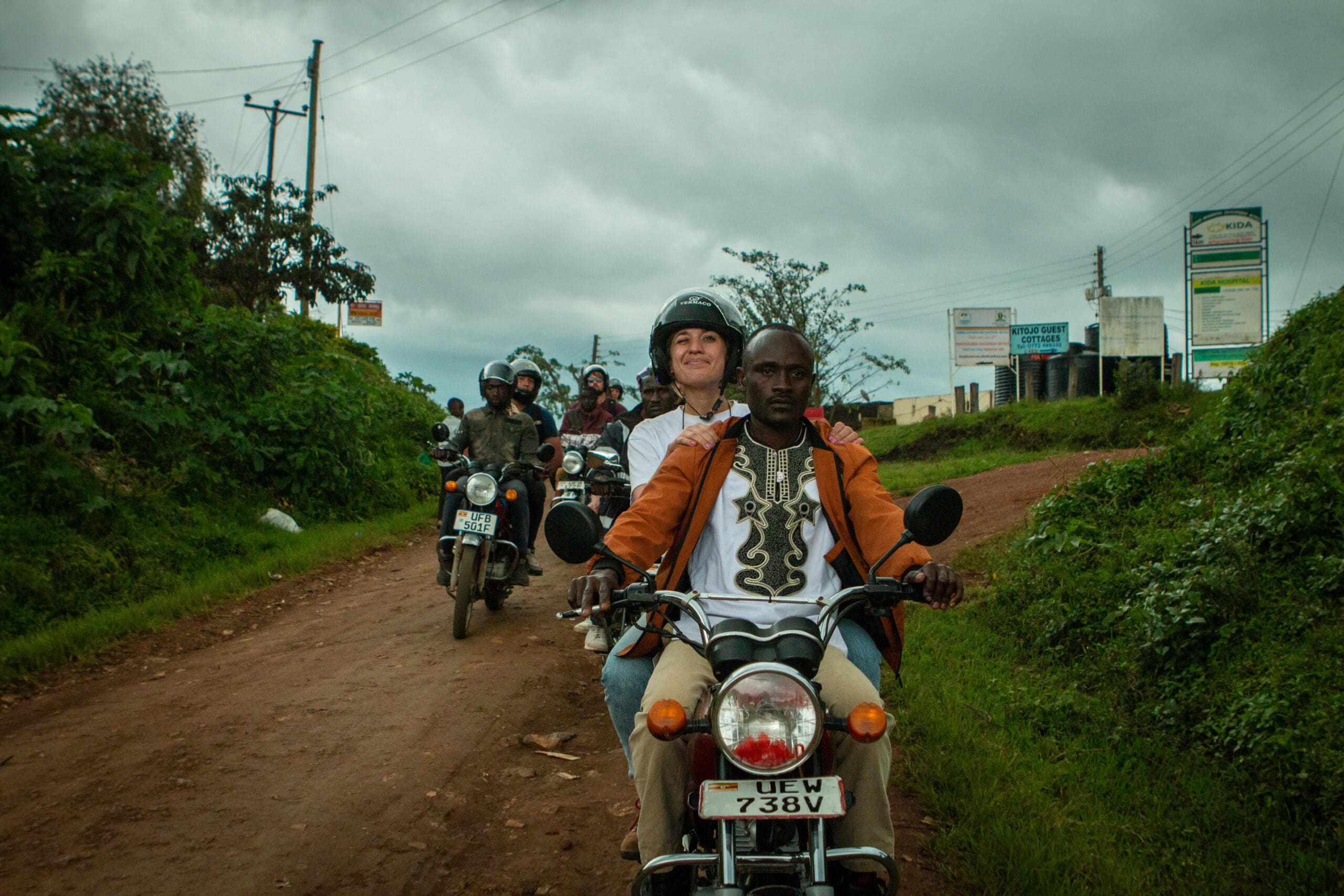 Een groep reizigers rijdt op een motor over een modderige plattelandsweg in Afrika, omgeven door groen onder een bewolkte hemel. De leidende motorfiets heeft een man aan het stuur en een lachende vrouwelijke passagier met een helm op. Andere motorrijders volgen.