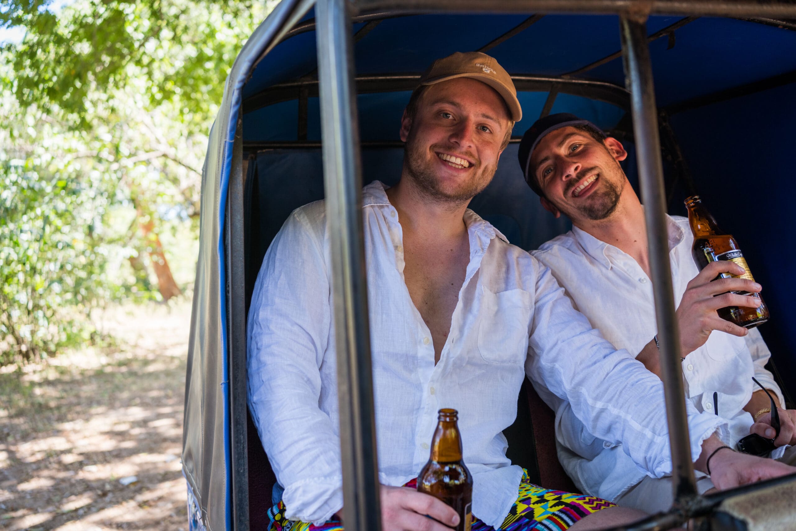Twee lachende mannen in witte shirts zitten ontspannen in een tuk-tuk, elk met een bierflesje in de hand. Zonlicht valt door de bomen op de achtergrond, wat de scène een zomerse, vrolijke sfeer geeft.