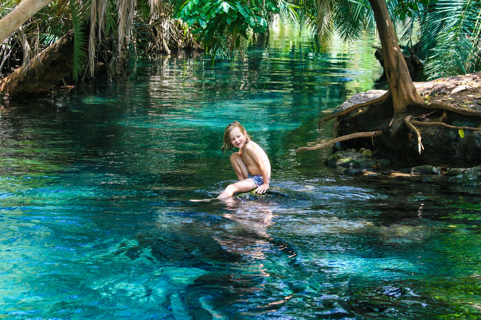 Een jong kind in zwemkleding zit en lacht op een rots in een helder, blauw natuurlijk zwembad omringd door weelderige groene bomen en planten - een beeld van de schoonheid van reizen door de prachtige landschappen van Afrika.
