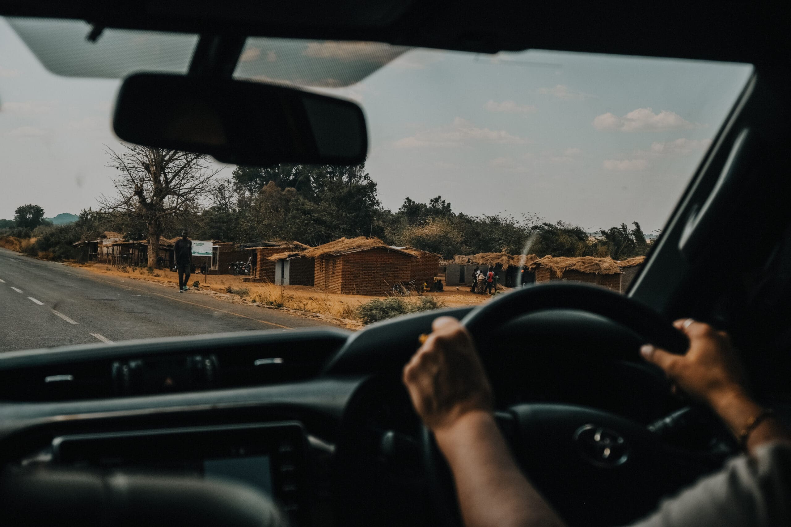 Een reiziger rijdt met een auto over een plattelandsweg in Afrika, langs huizen met rieten daken en mensen die langs het droge, zonnige landschap lopen. De foto is genomen vanuit de auto.