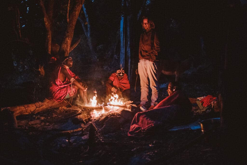 Verschillende reizigers in traditionele kleding zitten en liggen 's nachts rond een kampvuur in een Afrikaans bos. Het licht van het vuur verlicht hun gezichten en de nabijgelegen bomen, terwijl één persoon waakzaam in de buurt staat.