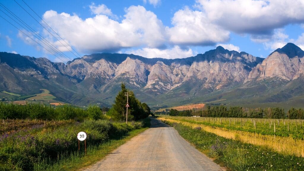 Een grindweg slingert door het Zuid-Afrikaanse boerenland in de richting van een bergketen onder een blauwe hemel met verspreide wolken. Velden en bomen omzomen de weg en leiden reizigers langs een bord met een maximumsnelheid van 30 km/u.