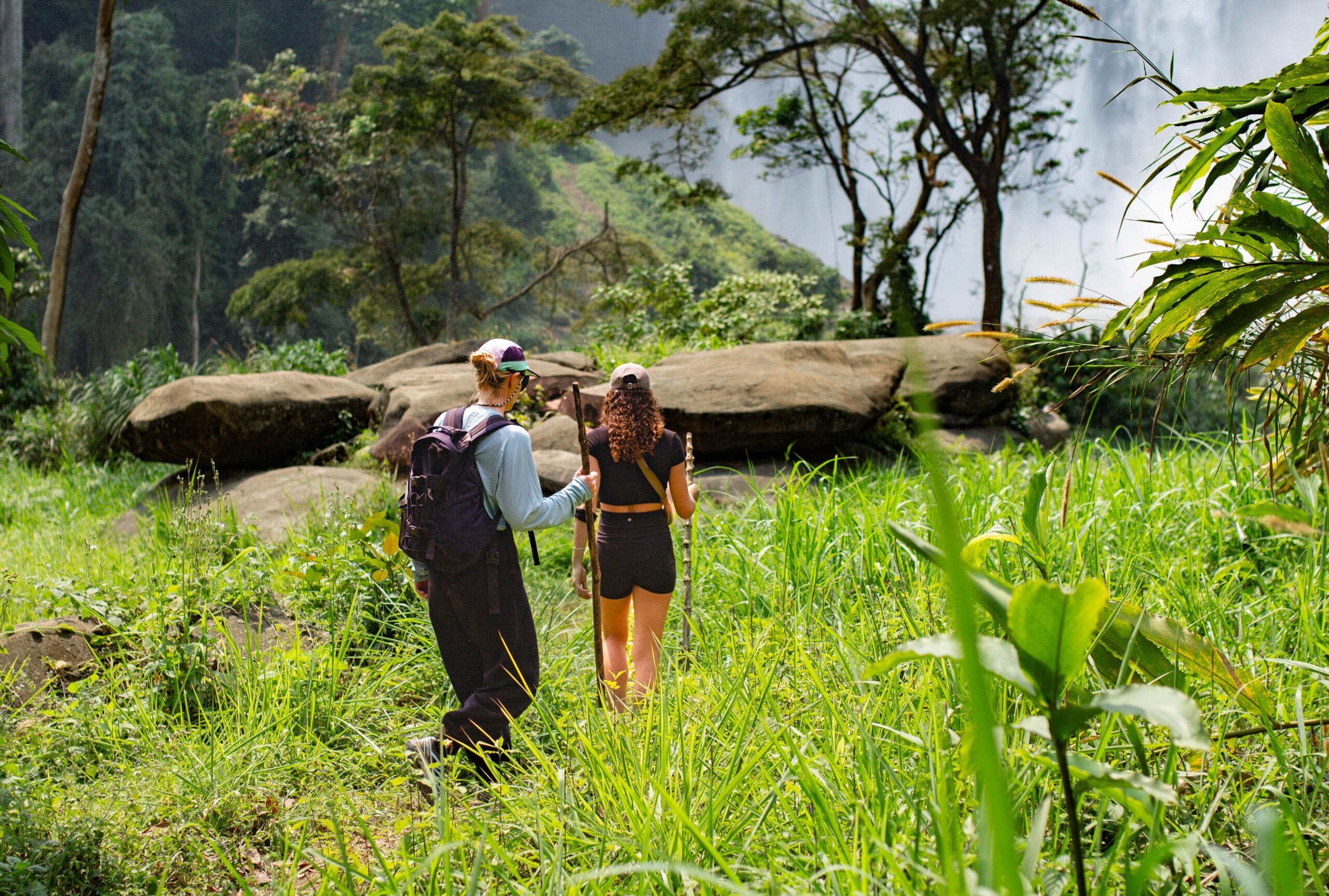 Twee reizigers met rugzakken lopen door weelderig groen gras in de richting van grote rotsen en bomen in Afrika, met een mistige waterval zichtbaar op de achtergrond.