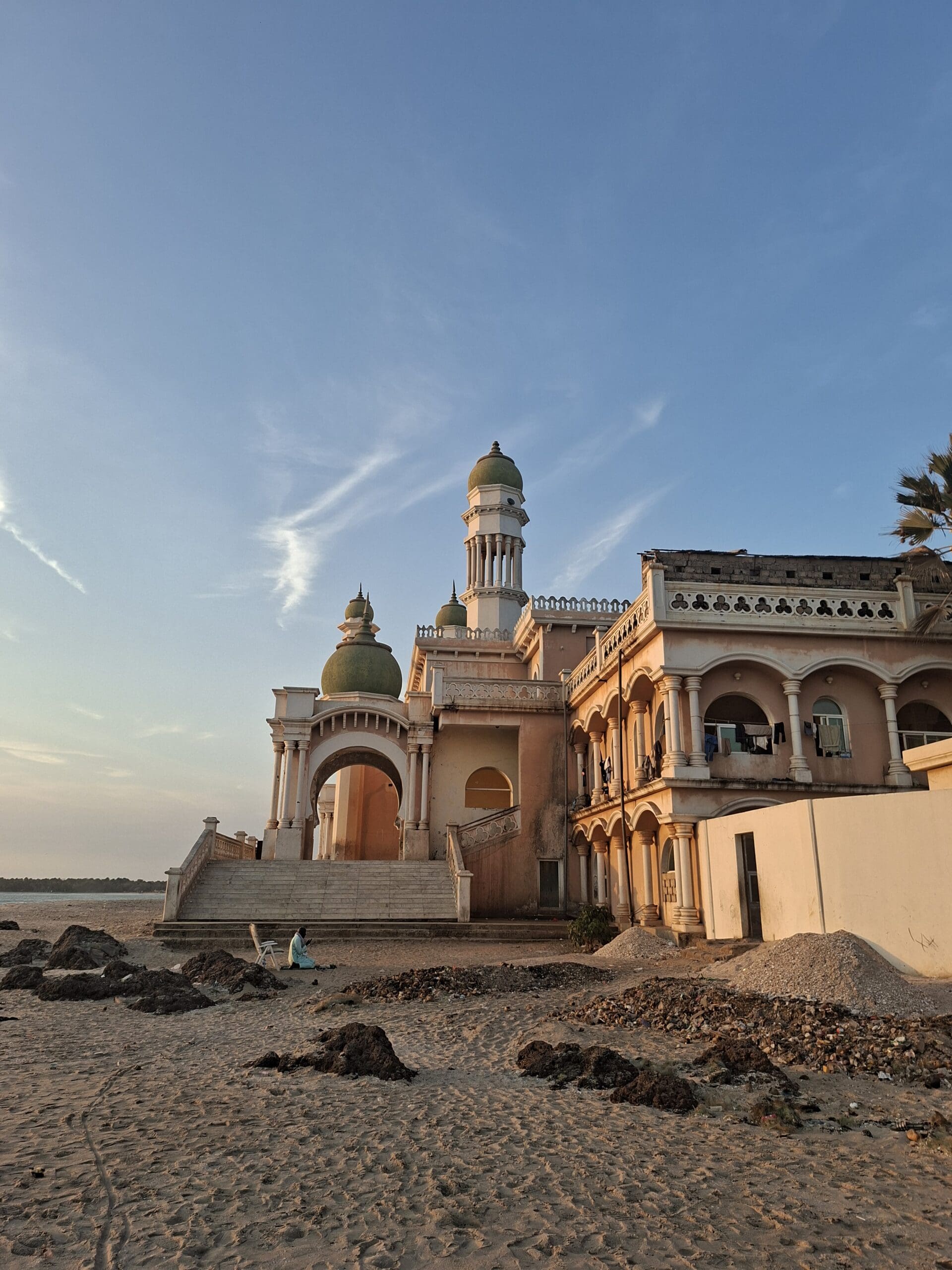 Ein prächtiges, helles Gebäude mit Kuppeln und Bögen steht bei Sonnenuntergang an einem Sandstrand; am Ufer sitzt eine Person, über ihr erstreckt sich ein strahlend blauer Himmel.