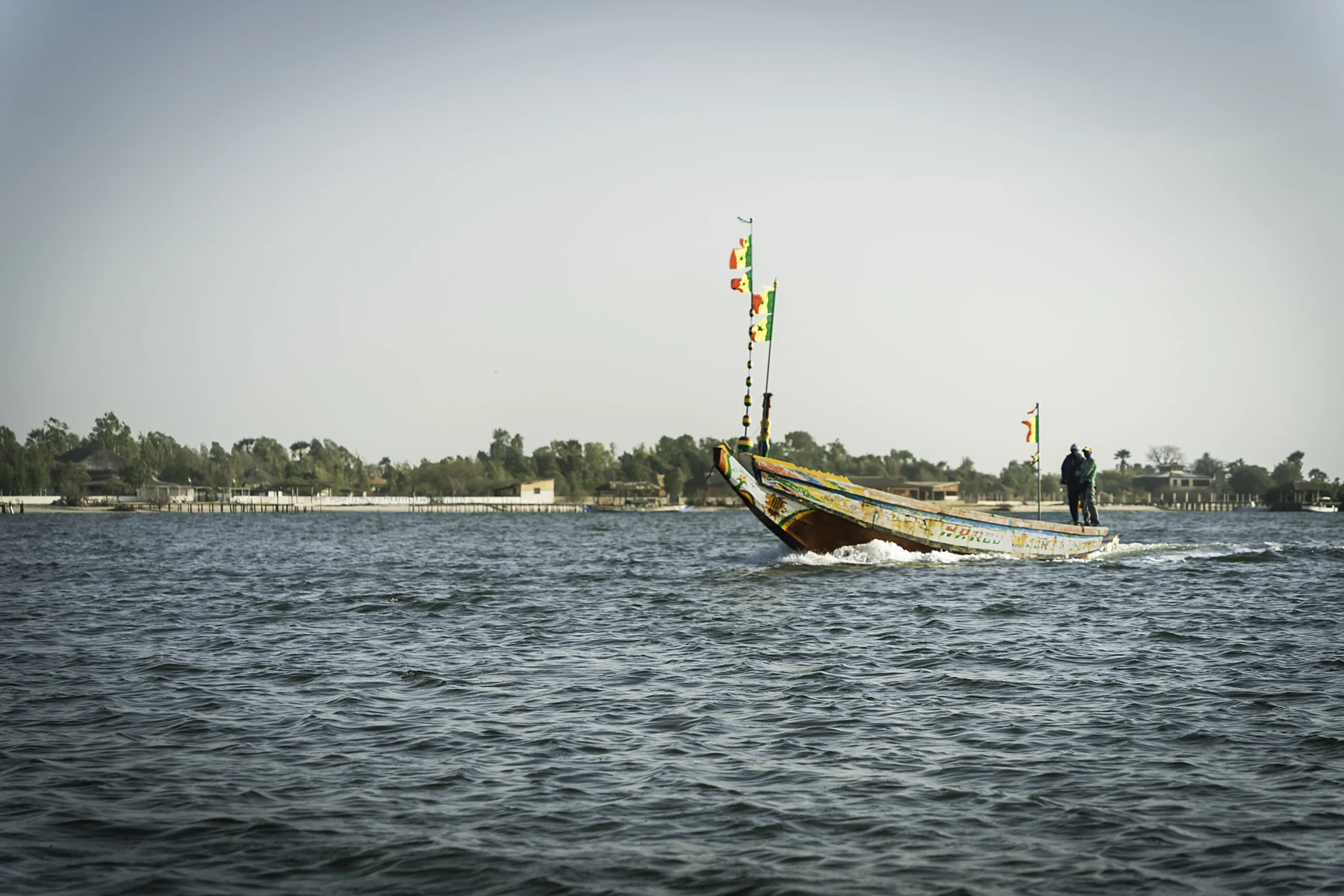 Ein buntes Holzboot mit Flaggen segelt auf einem ruhigen Gewässer; an Bord befinden sich zwei Personen, und in der Ferne ist unter einem dunstigen Himmel eine von Bäumen und Gebäuden gesäumte Küste zu sehen.