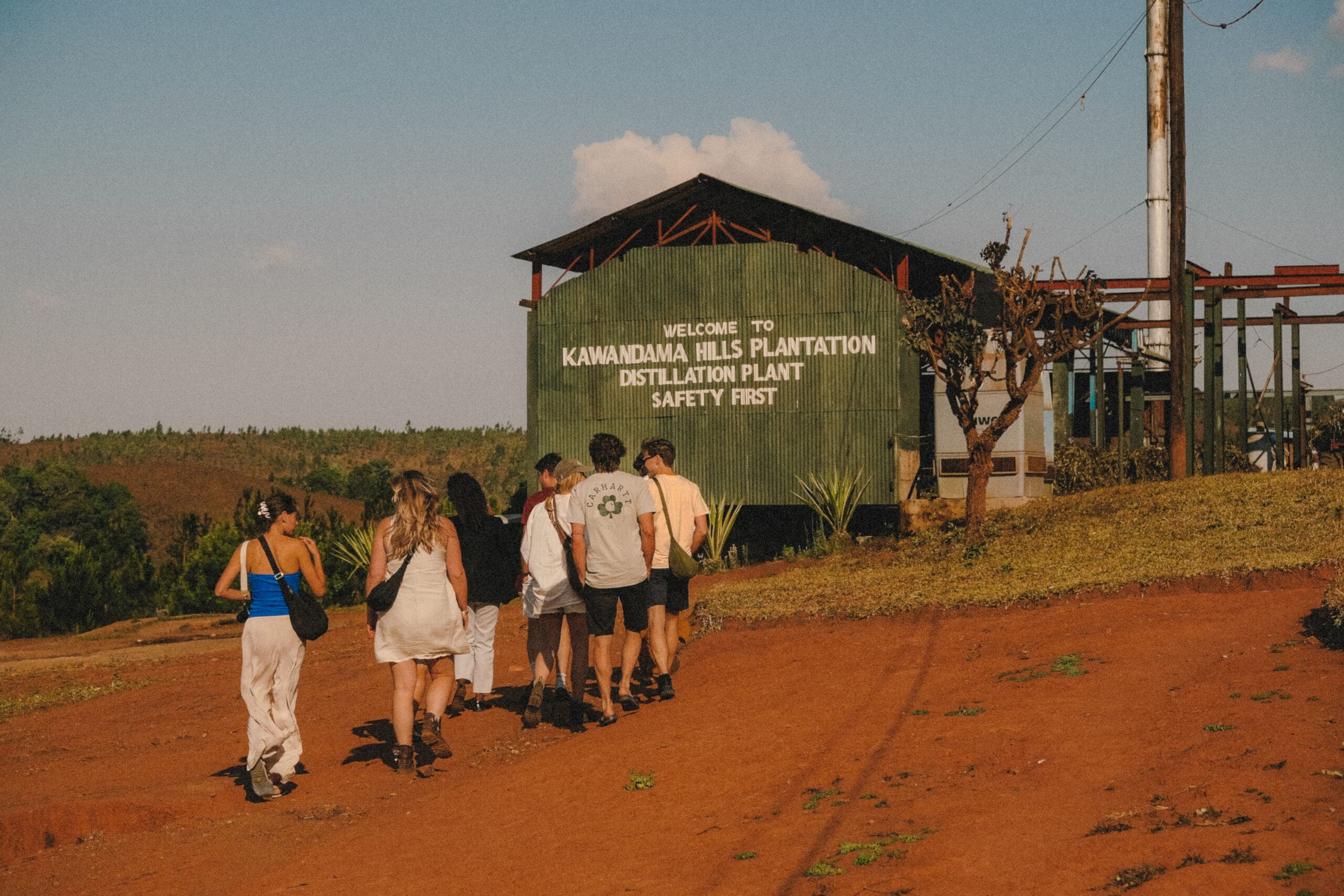 Een groep reizigers loopt over een rood zandpad onder de blauwe hemel van Malawi naar een groen gebouw met de tekst Welcome to Kawandama Hills Plantation Distillation Plant Safety First.