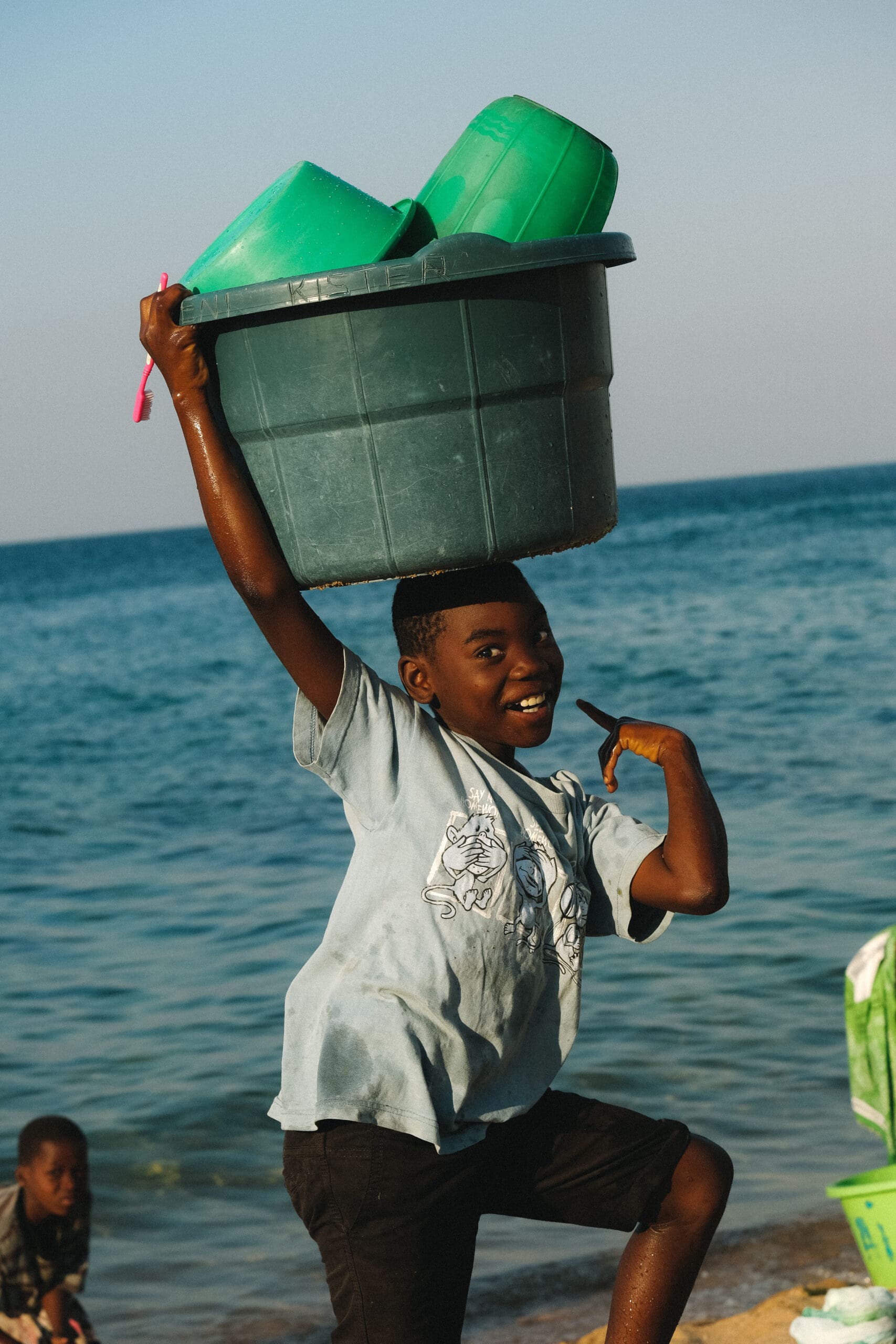 Een lachende jongen in Malawi staat bij het water en houdt een grote groene plastic bak vast met kleinere groene kommen op zijn hoofd. Hij wijst en poseert speels, terwijl een ander kind op de achtergrond bij de oever zit - een authentiek tafereel voor Afrika reizigers.