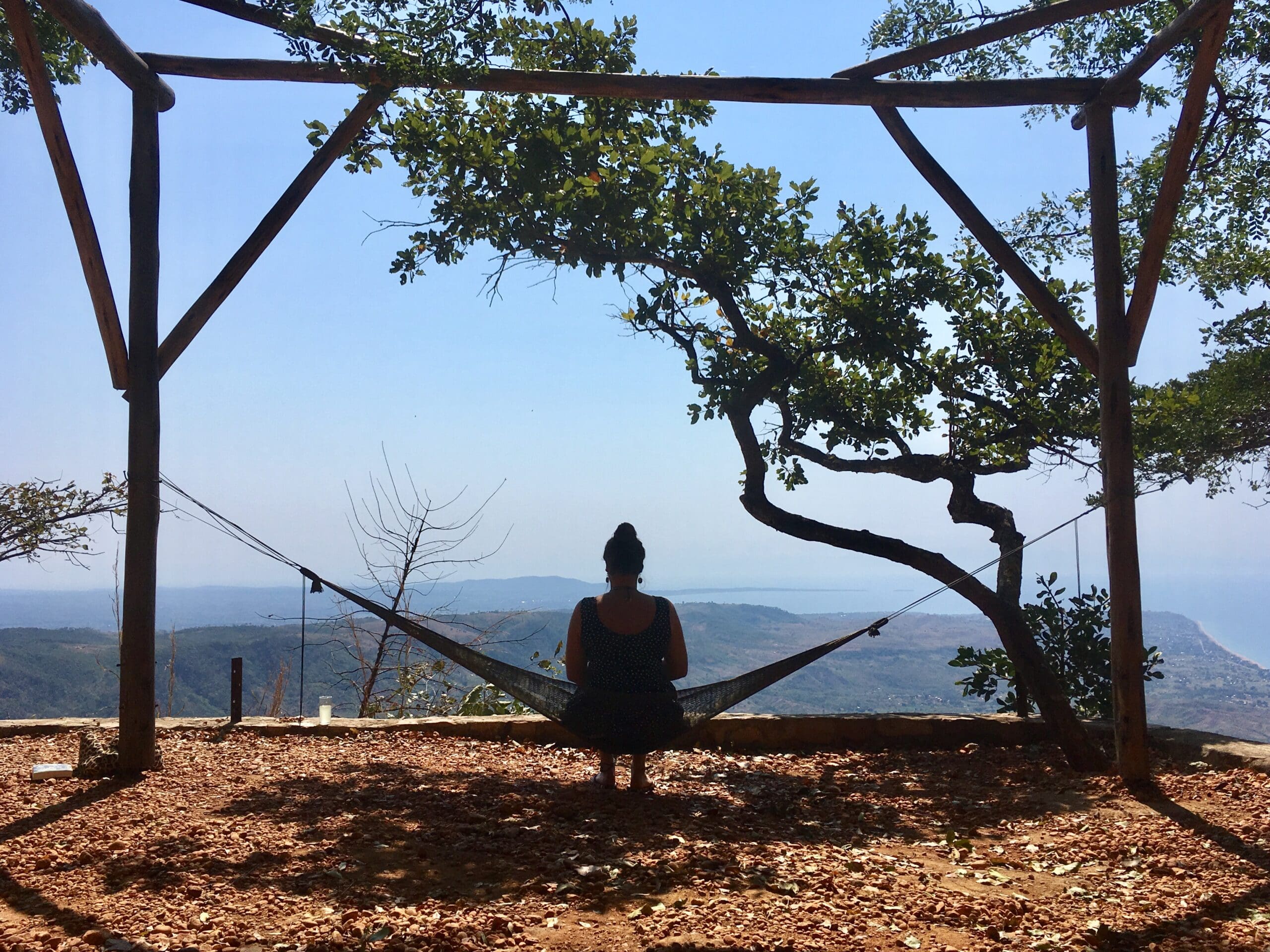 Een persoon zit in een hangmat onder een houten frame en boom, met uitzicht op een schilderachtig, bergachtig landschap van Malawi met een wazige lucht op de achtergrond.